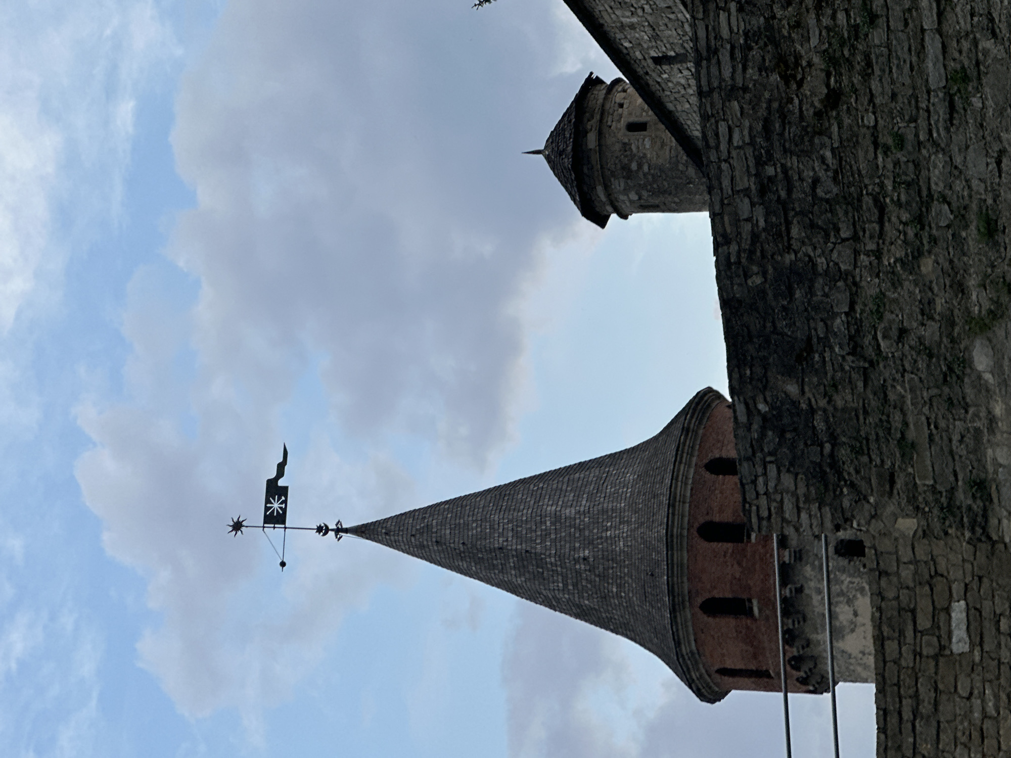 The image captures a view of a historic castle in Kamianets-Podilskyi, Ukraine, under a partly cloudy sky. The foreground is dominated by a rough stone wall. Behind this wall, a tall, conical tower with a weathered, shingled roof stands prominently. A weather vane with a star and a flag is visible at the apex of this tower. To the right, a shorter, cylindrical stone tower with a similarly shaped roof is partially visible, attached to a stone building. The architecture suggests a medieval fortress. No people or animals are present in the frame, and no specific actions or interactions are depicted. The environment is that of an outdoor, historical landmark.