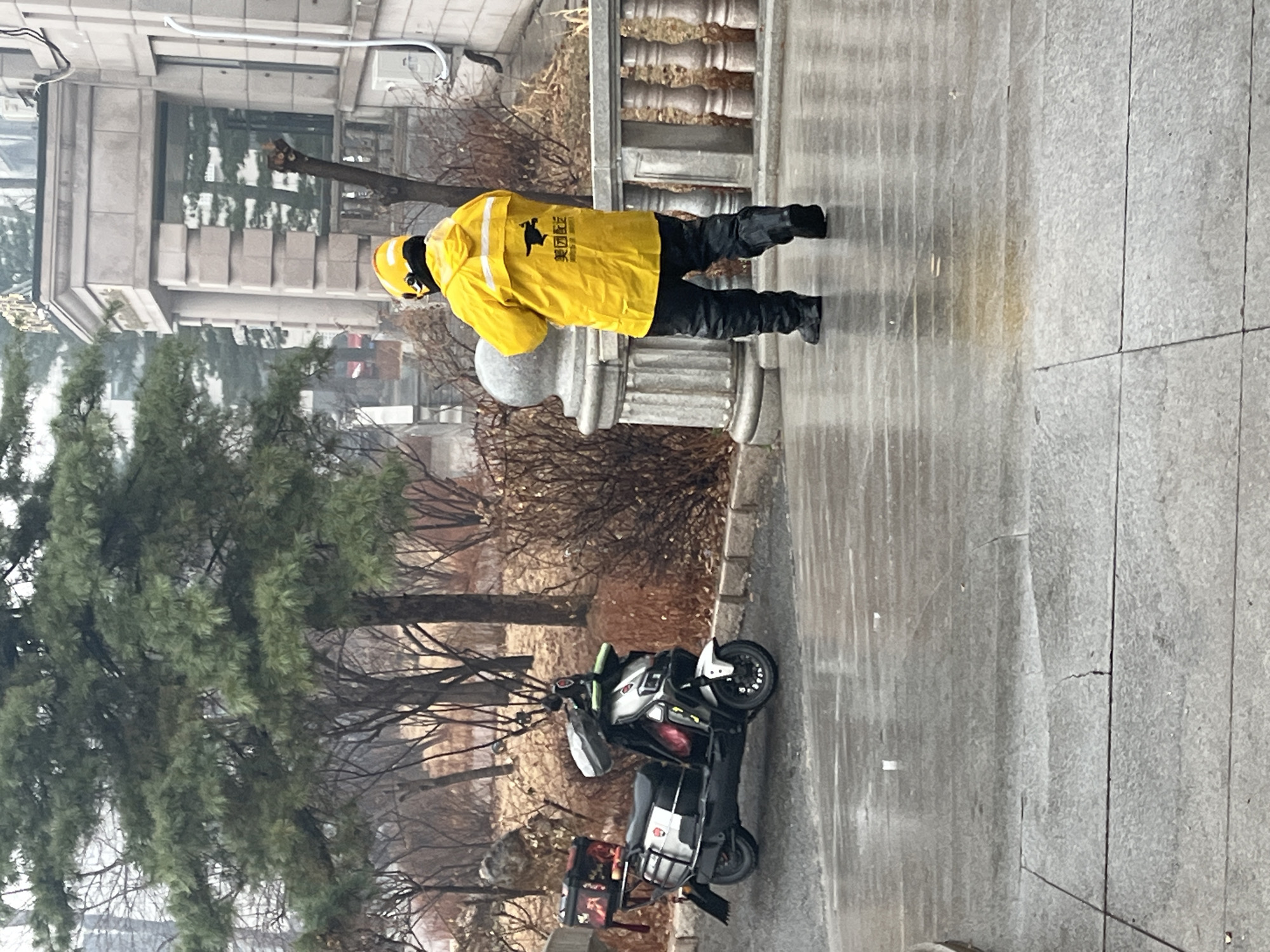 A delivery person is standing on a paved walkway in Changchun City, China. The delivery person is wearing a yellow jacket, black pants, and a yellow helmet. The delivery person is standing next to a column. A motorcycle is parked nearby with a delivery container affixed to the back. Trees are visible in the background, along with a building. The ground appears wet.