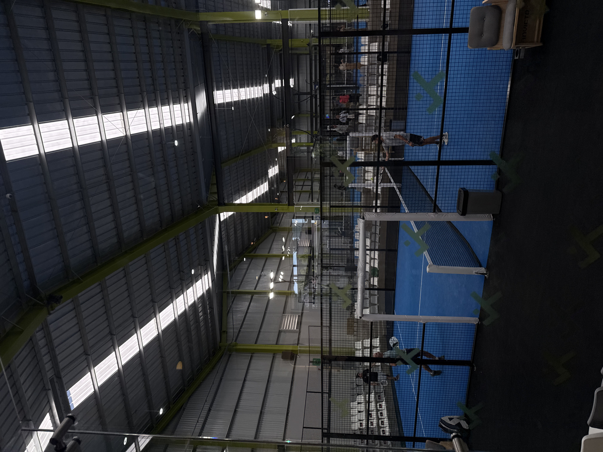 This media file captures an indoor padel tennis facility located in San Bernardino Tlaxcalancingo, Mexico. The scene features multiple padel courts with distinct blue playing surfaces marked by white lines, enclosed by black mesh and glass walls.
Several people are visible on the courts, actively engaged in playing padel or observing the games. They are holding padel rackets, indicating ongoing matches or practice sessions. The facility boasts a high, vaulted corrugated metal roof, interspersed with large, rectangular skylights that allow bright streaks of natural sunlight to illuminate the space. This suggests it is daytime. Supplemental artificial lighting fixtures are also present on the ceiling and along the yellow-green support beams.
Notable objects include white padel nets, a wooden bench with gray cushions, and a gray trash can placed next to one of the courts. A prominent stylized light green 'X' logo is painted on the blue court surfaces and occasionally on the mesh walls, serving as a recurring branding element. The word "INVICTO" is clearly visible on the side of one of the wooden benches. The overall atmosphere is one of active recreation within a modern, well-lit indoor sports complex.