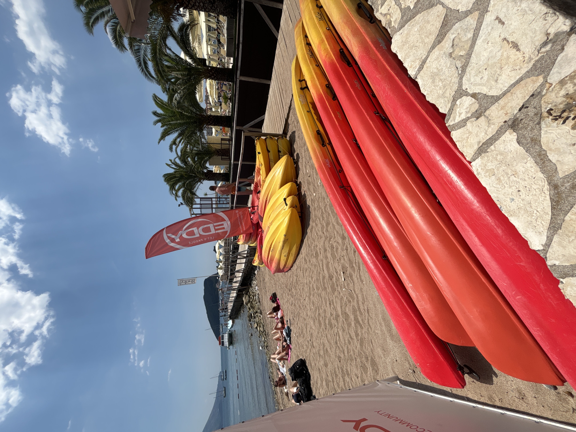 The image depicts a sunny day at a beach, likely in Budva, Montenegro, judging by the presence of kayaks and palm trees, common in coastal tourist areas. In the foreground, several red and orange kayaks are neatly arranged on the sand, bordering a decorative stone pathway. Further back, a group of people are sunbathing on towels on the sandy beach. A wooden pier extends into the calm, blue sea, where boats are visible. In the background, lush green palm trees line the shore, and there are beach umbrellas and lounge chairs set up, suggesting a resort or beach club. A red flag with the word "EDDY" prominently displayed is planted in the sand near the kayaks. The sky is a vibrant blue with scattered white clouds.