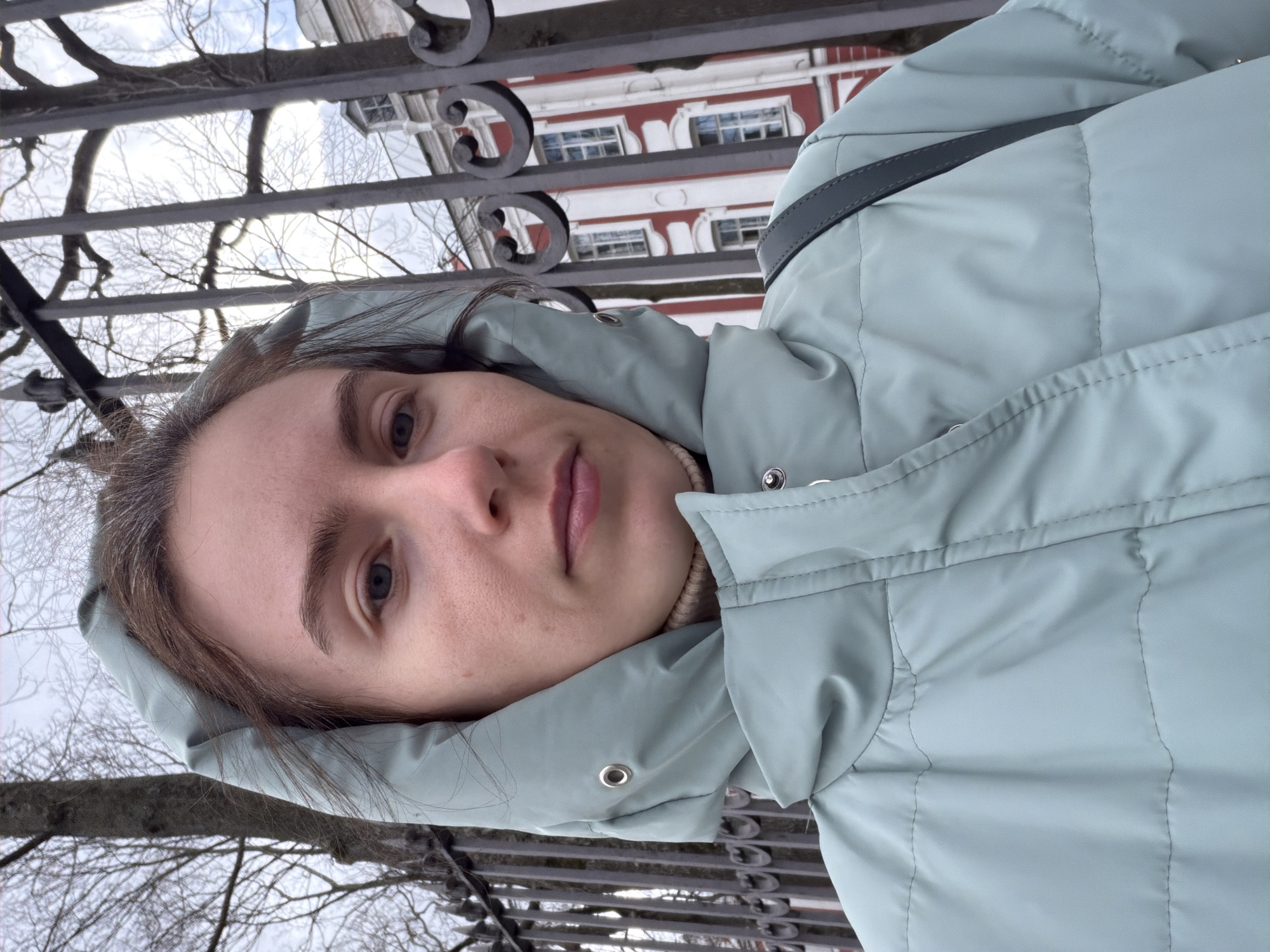 A young woman wearing a light blue hooded puffer jacket is positioned in front of a wrought-iron fence. The fence appears to be located in Saint Petersburg, Russia, judging from the architectural style of a building visible in the background. The building is multi-storied, with a red and white facade and symmetrically placed windows. The trees in the background are bare, suggesting it is winter. The woman is facing the camera. The image appears to be a selfie or a candid shot taken by another person.