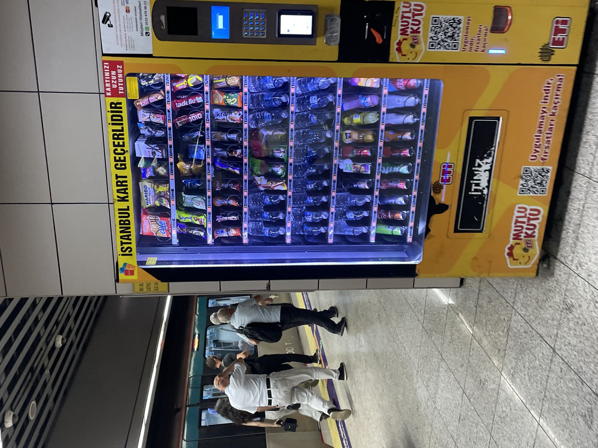 In this medium shot photograph, a bright yellow vending machine is the central focus, situated in a public transit station, likely a subway or metro. The machine is filled with various snacks and drinks, with labels and prices visible on some items. Above the snacks, a sign reads "ISTANBUL KART GEÇERLİDİR," indicating that the Istanbul Card is accepted. To the right of the vending machine, there is a payment terminal with a screen, keypad, and card reader. The floor is tiled with a light gray, speckled pattern, and a yellow line marks the edge of a platform. In the background, people are seen walking away from the camera towards a train, suggesting a busy urban environment. The overall lighting suggests an indoor, well-lit space typical of a transit hub.