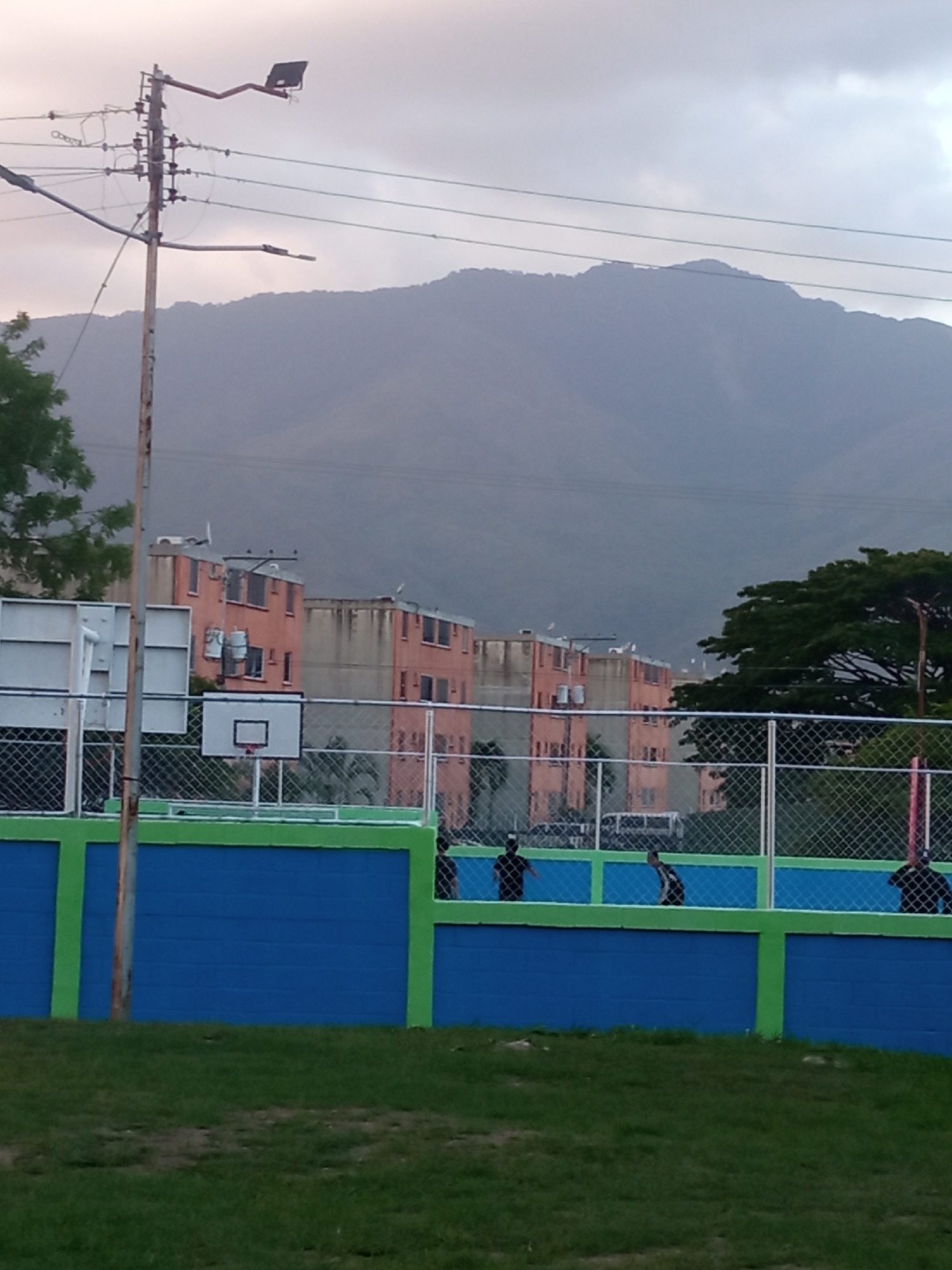 The image depicts an outdoor scene with a basketball court in the foreground. The court has a blue wall with green trim and a chain-link fence surrounding it. Several silhouetted figures, possibly people, are visible inside the court. Behind the court are multi-story apartment buildings with a peach-colored exterior. The backdrop features a large, misty mountain range under a cloudy sky. An utility pole with wires is on the left side of the frame. The location is likely within an urban setting near a mountainous area.