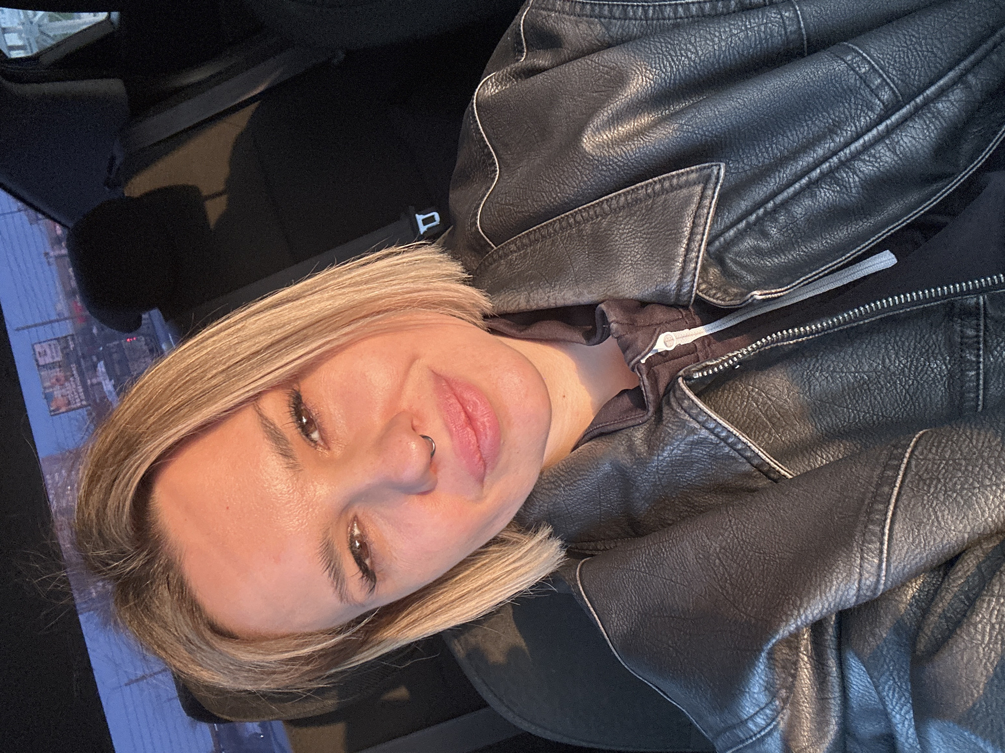 An individual, identified as female, is depicted in a close-up, selfie-style photograph taken inside a vehicle, presumably within Rostov-on-Don, Russia. The subject has short, light blonde hair and brown eyes, facing the camera with a subtle smile. A silver-colored septum piercing is visible. She wears a black faux leather jacket with white stitching details on the lapels and shoulders, over what appears to be a dark, partially unzipped hoodie with a light grey zipper pull. Warm, natural light illuminates the right side of her face (viewer's left). The background shows the dark interior of a vehicle, with a black headrest and a seatbelt buckle receiver visible behind her. Through the vehicle's rear window, a blurry urban environment is discernible, consistent with a city setting like Rostov-on-Don, featuring indistinct buildings, structures, and at least two advertising billboards. The ambient lighting suggests either late afternoon or early morning conditions.