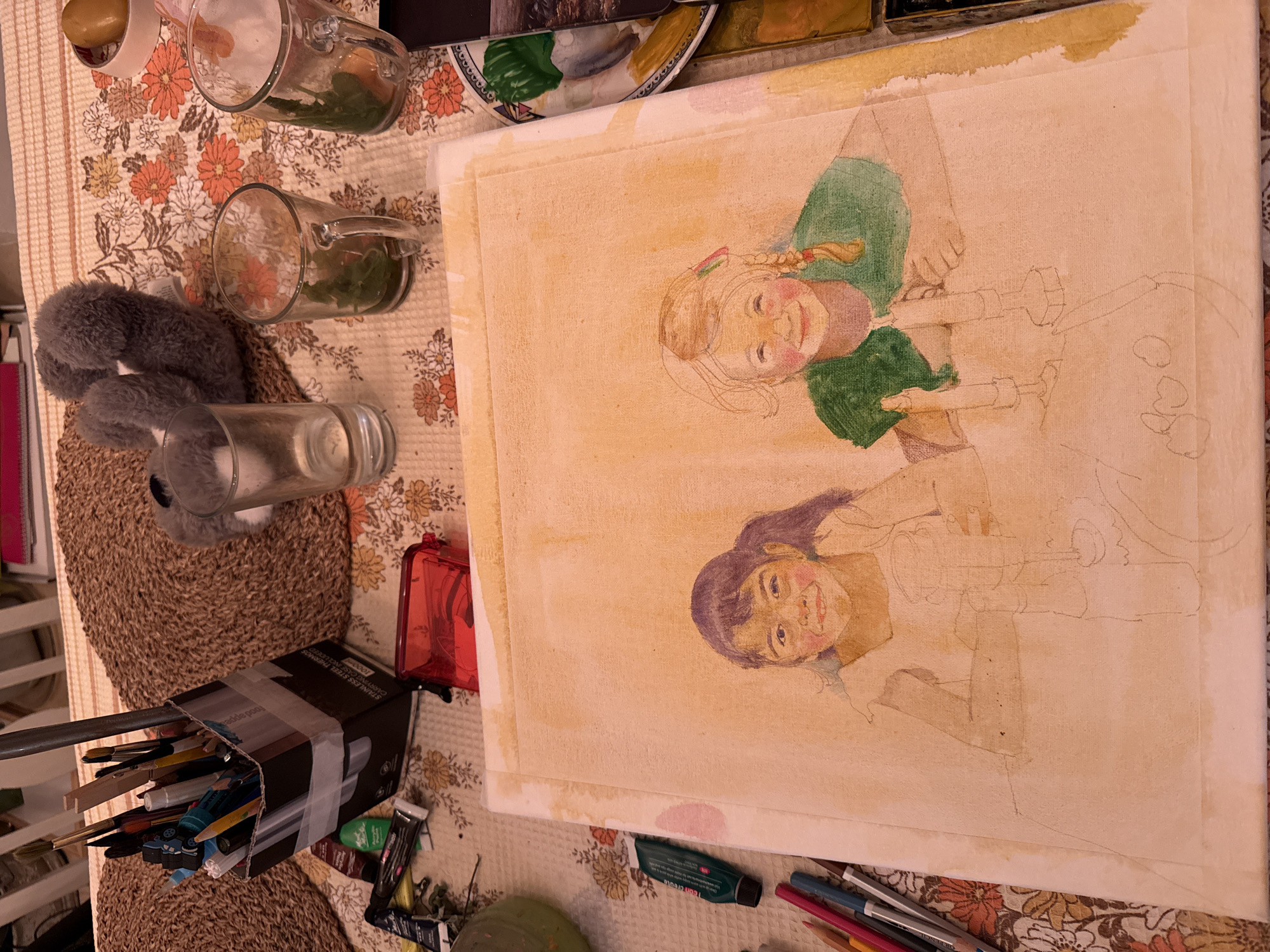 The image depicts an art studio setting on a table. A partially completed watercolor painting of two young girls is the central focus. The girl on the left has dark hair in a ponytail and is wearing a light-colored shirt, with her face partially rendered. The girl on the right has blonde hair with braided pigtails and is wearing a green shirt, her face also partially completed. Both girls have rosy cheeks. The painting is on a canvas, with faint pencil lines visible for the rest of the composition, which appears to include candlesticks and potentially other objects on a table.
Scattered around the painting are various art supplies. To the left, a collection of paint tubes, including a green tube, and colored pencils are visible. In the background, a cardboard container holds an assortment of paintbrushes and pens. Several drinking glasses, some containing what appears to be greenery, are also on the table, along with a red pencil sharpener. A small, gray plush toy is visible behind the glasses. The table is covered with a floral-patterned tablecloth, and a woven placemat is partially visible. The scene appears to be indoors, possibly in a home studio in Kfar Vradim, Israel.