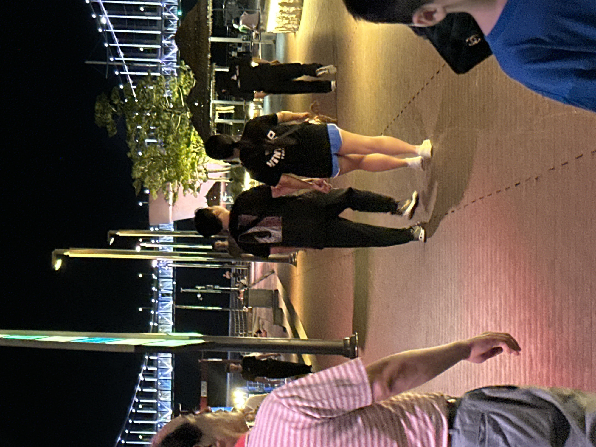 The image depicts a nighttime scene on a pedestrian walkway, possibly along the coast of Busan, South Korea. In the foreground, a person from the left side of the frame, wearing a pink and white striped shirt and gray pants, is walking away from the camera. To the right of this individual, a couple is walking hand-in-hand. The man is wearing a black t-shirt and black pants, while the woman is wearing a black t-shirt and blue shorts. Behind them, other individuals are visible on the walkway, some walking and some standing. In the background, a brightly lit bridge, potentially part of the Busan Harbor Bridge or a similar coastal structure, spans across the water. The environment appears to be an urban waterfront area, with decorative lighting, trees, and covered seating areas visible. The overall impression is a lively evening atmosphere with people enjoying a stroll.