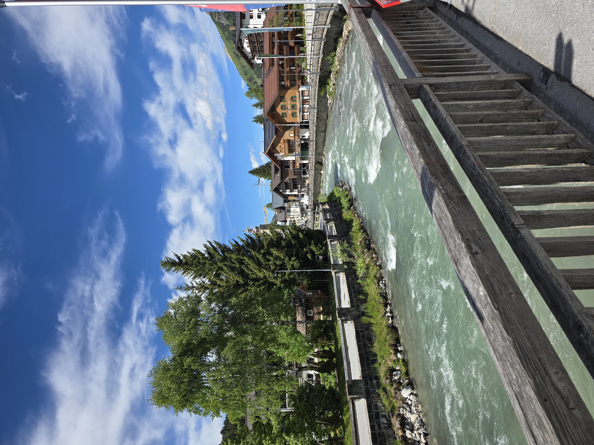 This is a full-frame, eye-level, outdoor, vertical shot of the Lech river in Lech, Austria. The river flows from left to right across the frame, with white water evident in its turbulent flow. On the left bank of the river, a stone wall with a sidewalk above it runs parallel to the water. Behind the sidewalk, a cluster of trees and buildings is visible. On the right bank, a wooden railing forms the foreground, and beyond it, a street and a row of buildings, characteristic of alpine architecture, are situated. These buildings feature wooden facades, balconies, and pitched roofs, with some having awnings. A pedestrian bridge spans the river in the middle ground. The sky is a vibrant blue with scattered white clouds. The lighting suggests it is daytime, likely late morning or early afternoon. No people are visible in the scene.