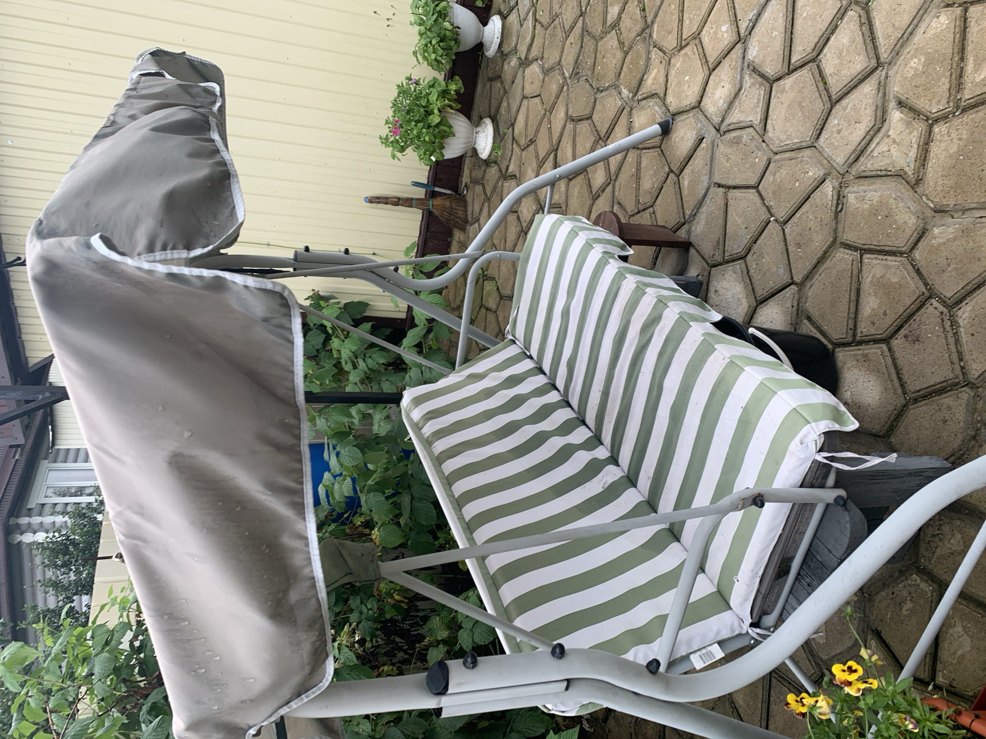 The image shows a garden swing set with a green and white striped cushion. The swing set has a beige canopy that appears to be wet, suggesting it may have recently rained. The swing is on a paved patio made of interlocking hexagonal stones. In the background, there is a light yellow wooden fence, a log house, and some greenery. Two white planters with pink and green flowers are also visible. A broom leans against the fence. There are no people in the image. The scene is outdoors in what appears to be a residential backyard.