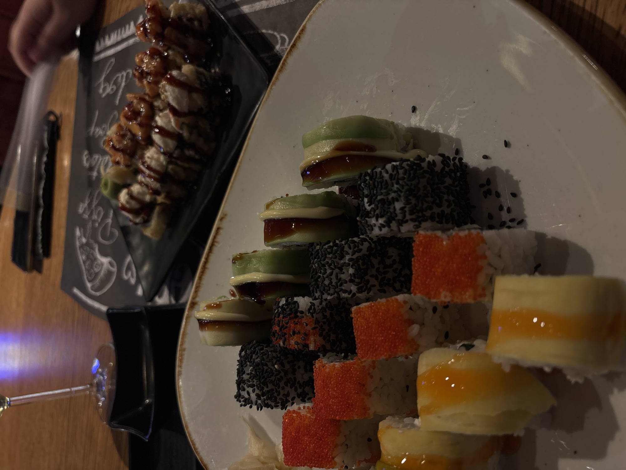 The image depicts two plates of sushi on a wooden table. One plate is black and contains sushi rolls topped with a dark sauce. The other plate is white and has a stack of sushi rolls decorated with avocado, black sesame seeds, orange tobiko, and yellow mango. A wine glass is visible in the lower-left corner. The setting appears to be a restaurant. There is no way to locate the specific restaurant or location in Varna, Bulgaria. No people or actions can be determined from the image.