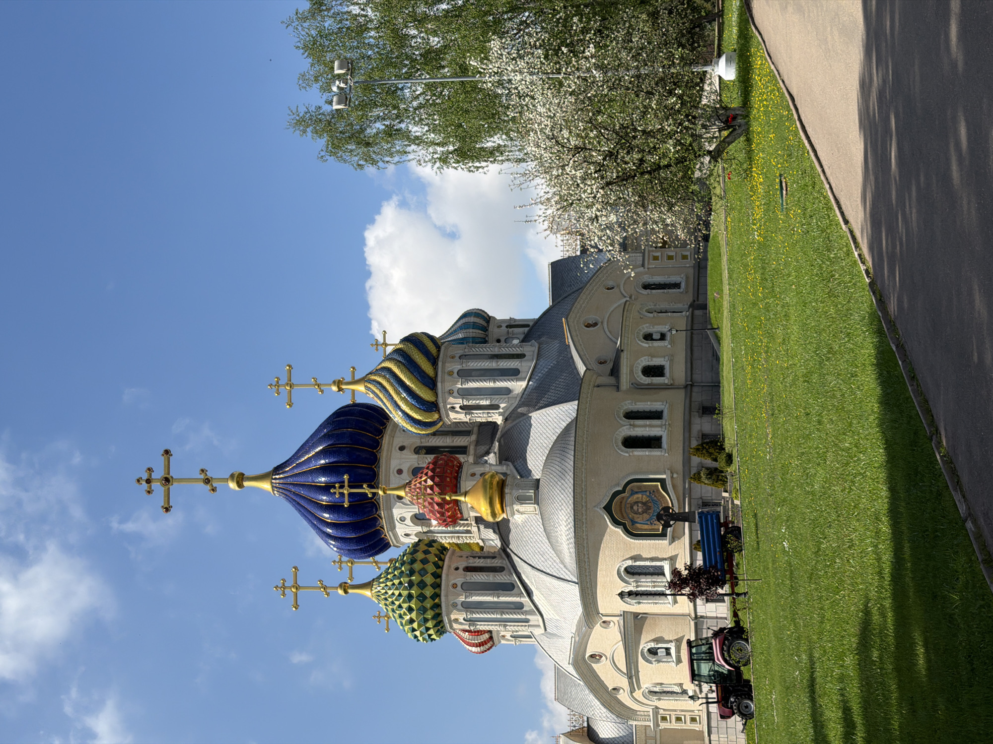 The image depicts the Izmailovo Kremlin in Moscow, Russia, showcasing a colorful architectural ensemble dominated by onion-shaped domes characteristic of Russian Orthodox churches. Three prominent domes, colored blue, gold/red, and green, are visible against a partly cloudy blue sky. The building itself features ornate detailing, arched windows, and mosaic artwork. To the right of the church, a green lawn stretches towards a paved walkway. A street lamp and flowering trees are also visible. A tractor is parked on the lawn.