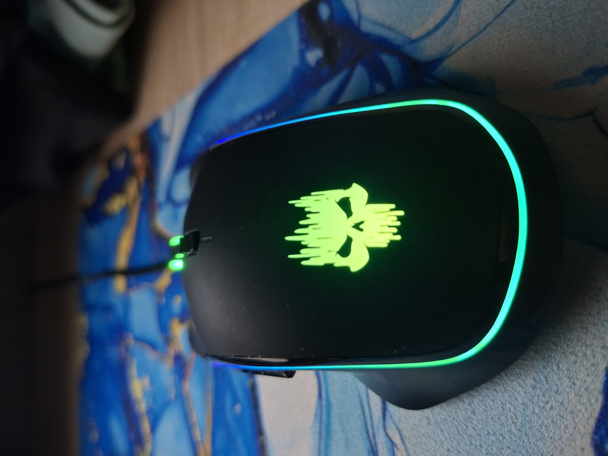 A black computer mouse sits on a mousepad featuring a blue and gold marbled pattern. The mouse has a glowing green skull design in the center and a rim of RGB lighting around its edge. The cord leading from the mouse is visible on the left. In the background, there is a blurry object that looks like a mug.