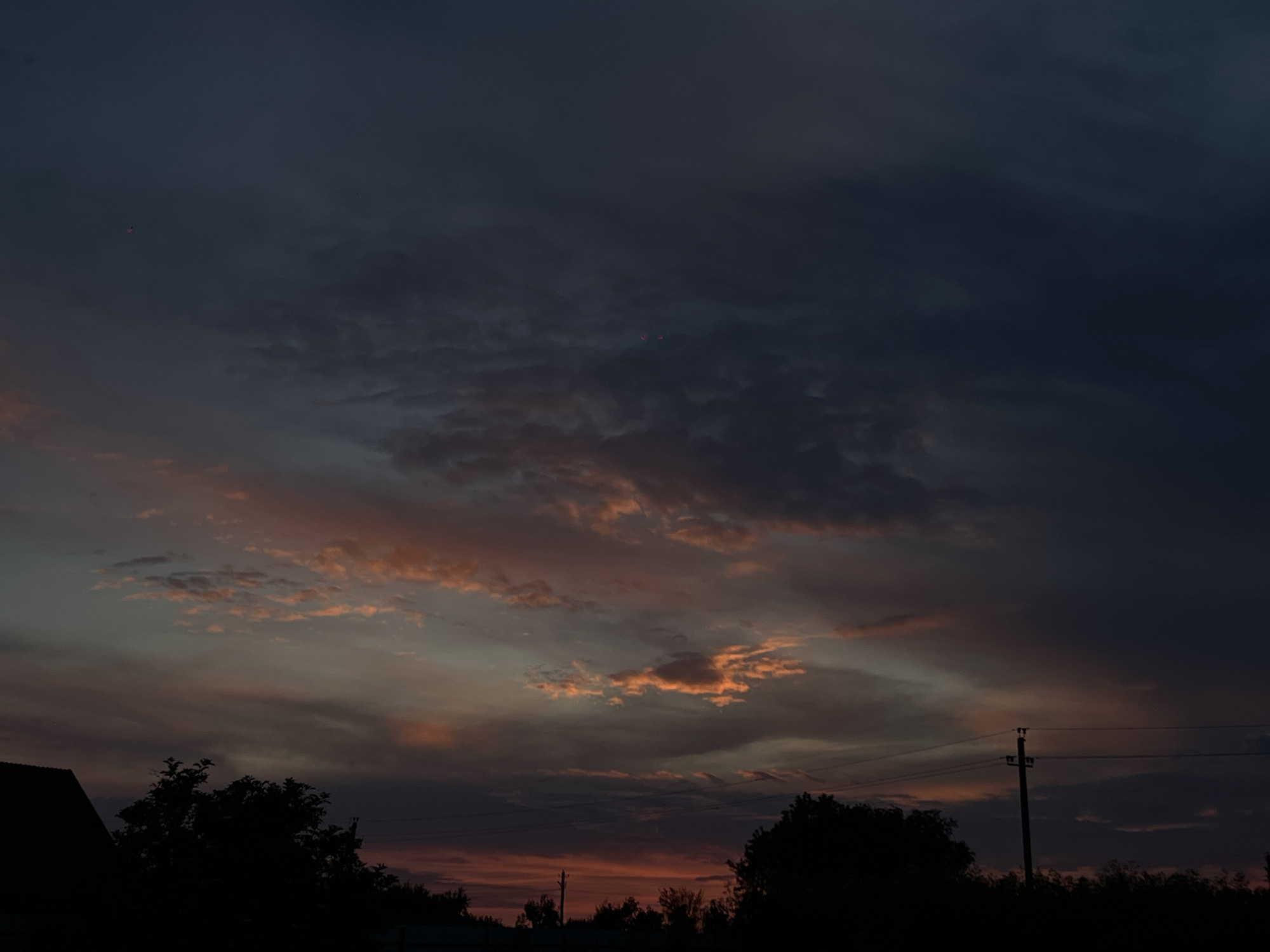 The image shows a dark, cloudy sky at dusk, with hints of orange and pink light peeking through the clouds. Silhouettes of trees and a building are visible against the horizon. A utility pole with power lines is also present. The scene is consistent with a sunset or twilight in Novopokrovskaya, Russia.