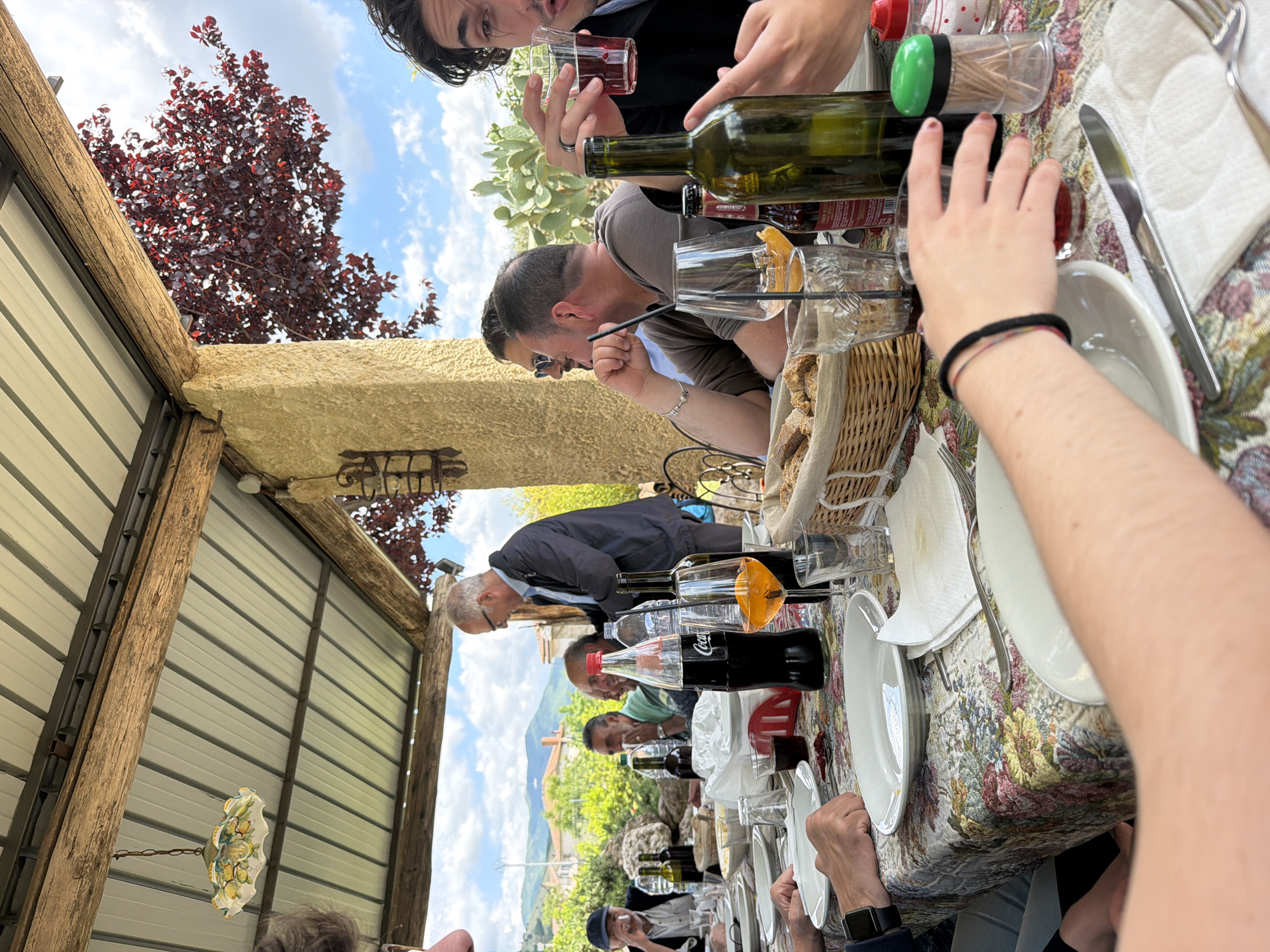 A daytime view shows a group of people gathered at a long table outdoors in Sala Consilina, Italy. The table is covered with a floral patterned tablecloth and set with white plates, silverware, and glasses. There are bottles of wine, a Coca-Cola bottle, and a bread basket on the table. People are seen seated around the table, engaging in conversation and drinking. The perspective is at an angle, with part of the view including a tree with red foliage and a structure with a metal roof and wooden beams. The sky is visible in the background.
