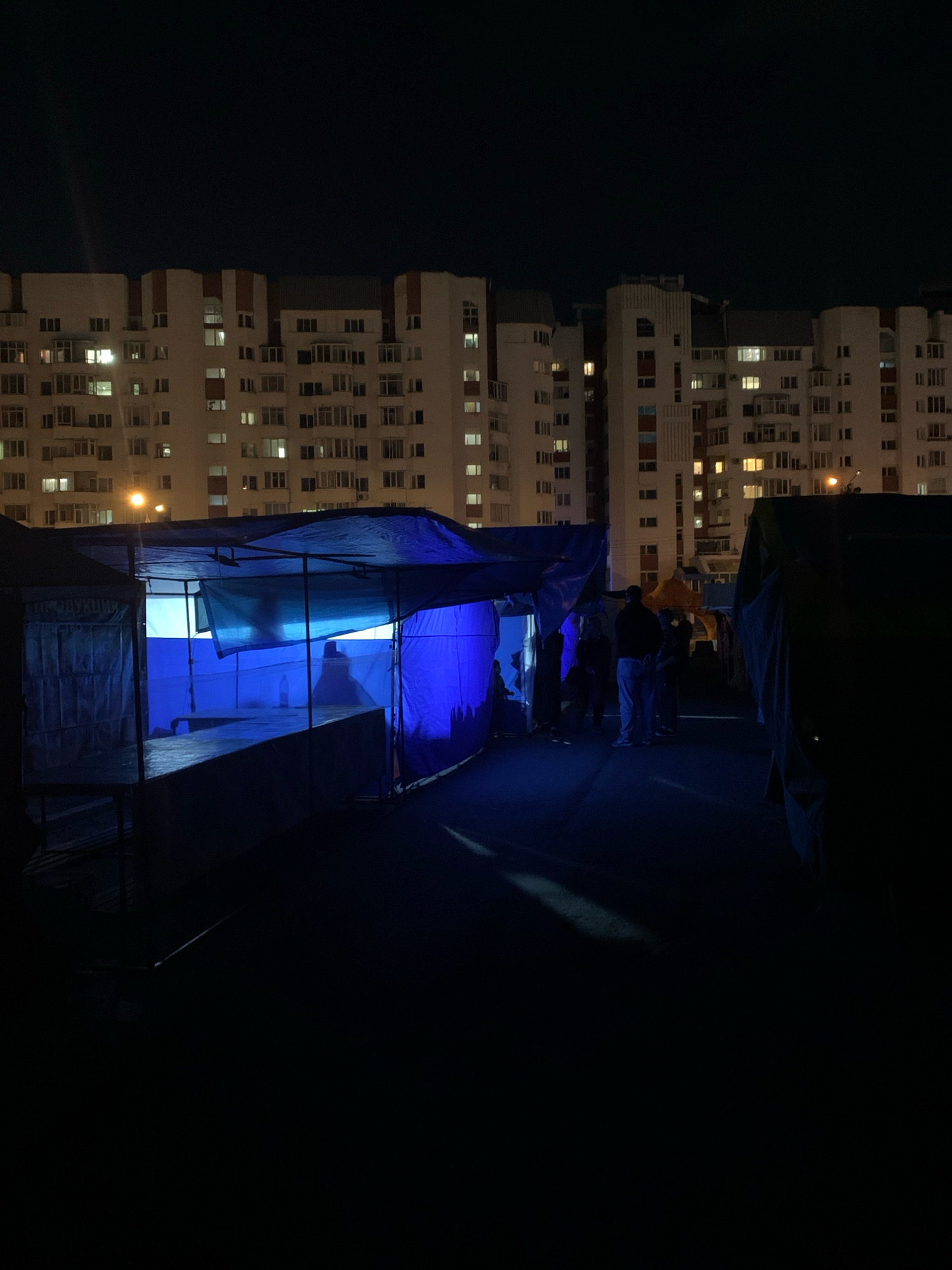 The image depicts a nighttime street scene in Arkhangelsk, Russia, with several temporary market stalls set up in front of a large, illuminated apartment building. The stalls are covered with blue tarps, creating a vibrant, artificial light. In the foreground, a stall is partially visible, with its interior lit by a bright blue light. A shadowy silhouette of a person is visible inside this stall.
Further back, a group of people are gathered near another stall. Some are standing and talking, while others appear to be browsing. The apartment building behind the stalls is brightly lit, with many windows illuminated, suggesting it is residential. The overall atmosphere is that of a nighttime market or gathering, with the stark contrast between the artificial light of the stalls and the residential glow of the building. The ground is dark and paved, possibly asphalt. The sky is a deep, dark blue, indicating it is late at night.