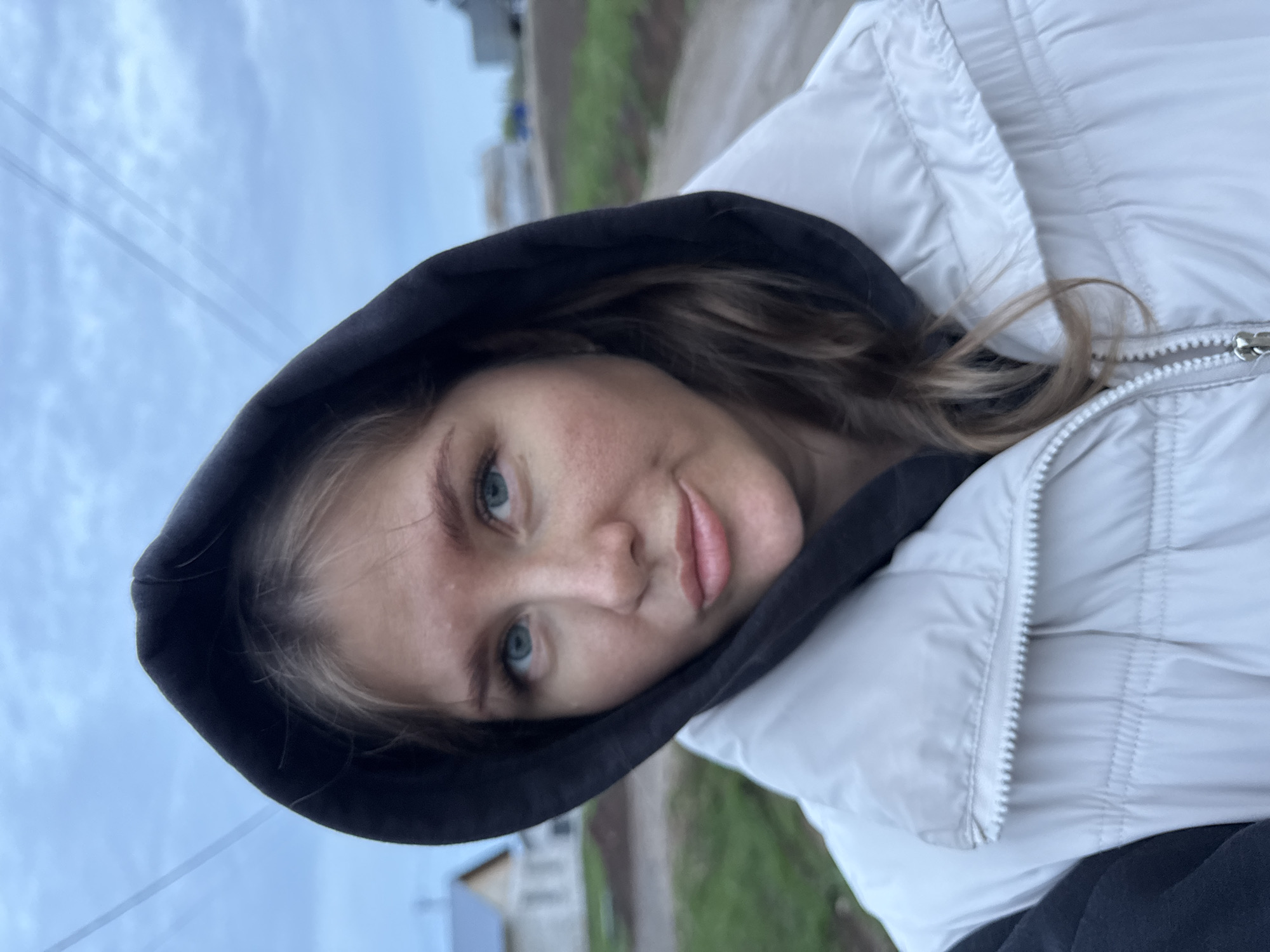 A selfie of a young woman with fair skin and light brown hair, wearing a black hoodie under a white puffer jacket. She has blue eyes and is looking directly at the camera with a slight smile. The background appears to be an outdoor environment, likely in a residential area of Ферма, Russia, with green foliage and buildings visible in the distance. The sky is overcast.
