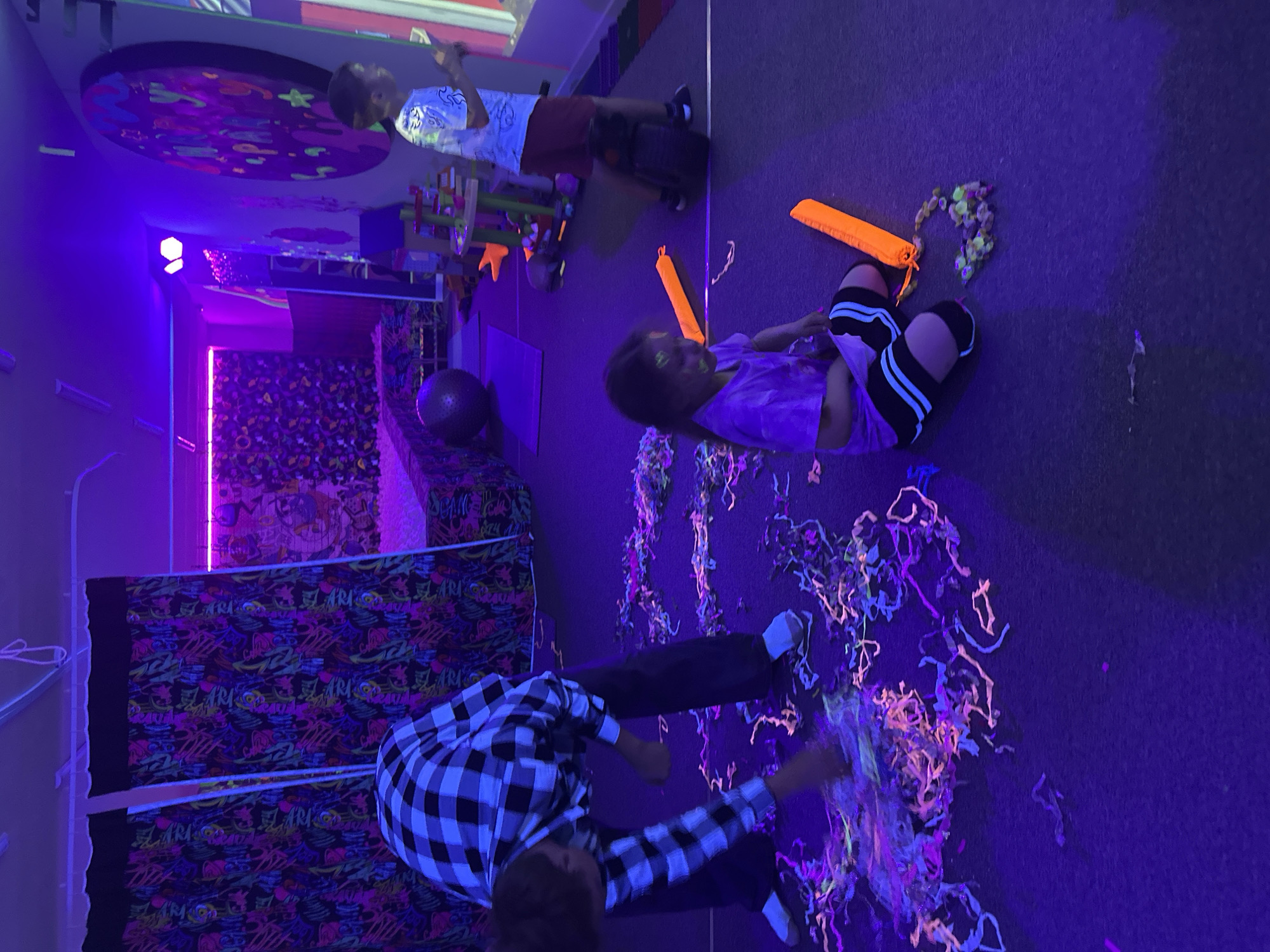 The scene shows three children indoors under ultraviolet or blacklights. The setting appears to be a recreational or entertainment facility. One child in a black-and-white plaid shirt is interacting with fluorescent streamers on the floor. Another child is sitting on the floor near a drawstring bag and scattered objects; they wear a purple shirt and striped leggings. A third child, wearing a white shirt and maroon shorts, stands further back in the room. A portion of a wall or curtain in the background features a colorful graffiti design, and there are various toys and play structures visible. The location is Unknown, Unknown.