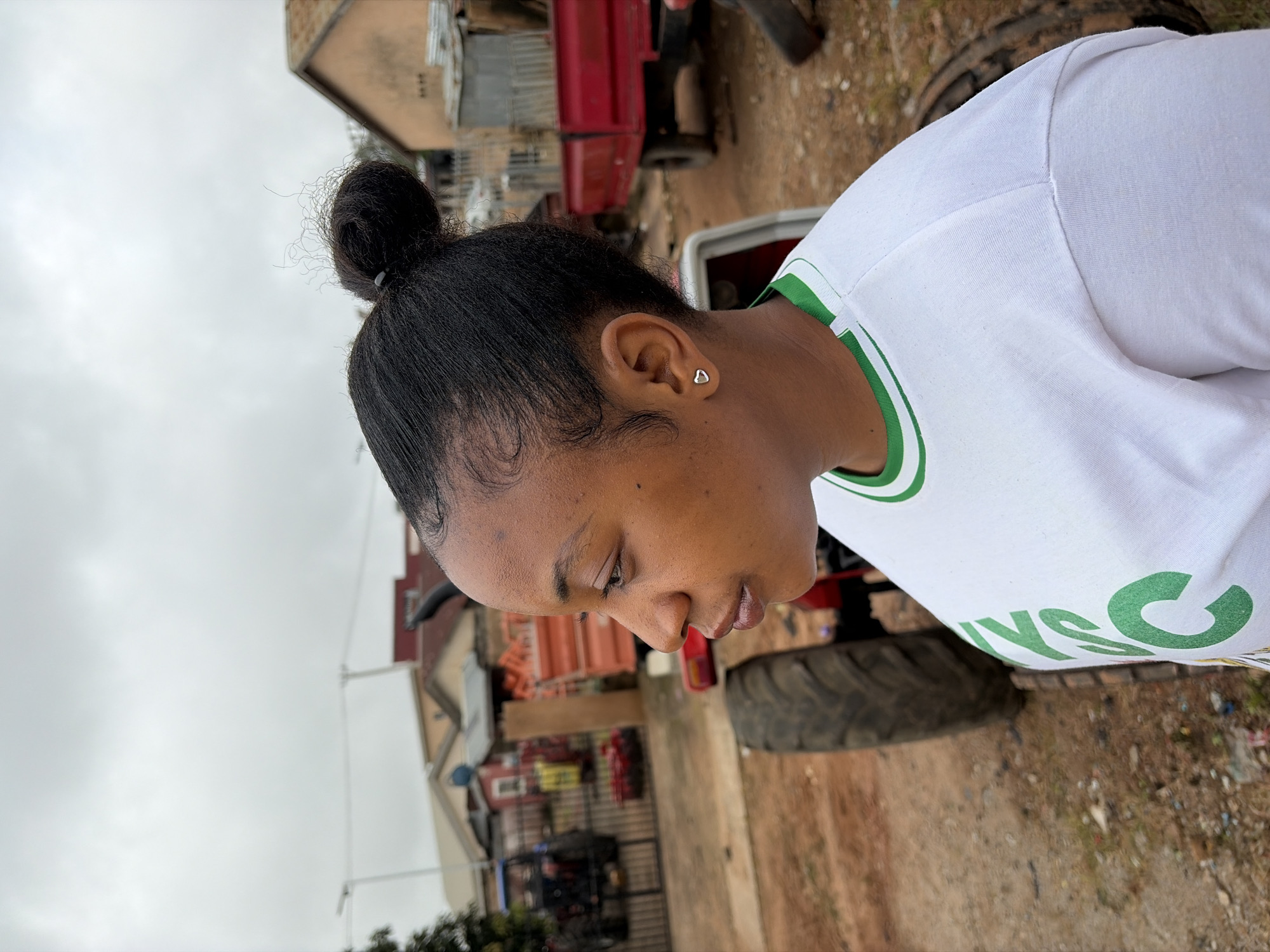 This is a medium shot, profile image of a young Black woman with her hair pulled up in a bun. She is wearing a white t-shirt with green trim and green lettering on the front that appears to read "NYSC". She has a small silver heart-shaped earring in her left ear. She is looking to her left, with her gaze directed towards something outside the frame.
The background suggests an outdoor setting in a semi-rural or peri-urban area. In the middle ground, there are several agricultural vehicles, including a red tractor and a red trailer. Beyond the vehicles, there are buildings that appear to be residential or small commercial structures, some with corrugated metal roofs. The ground is unpaved and appears to be dirt and gravel, with some scattered debris. The sky is overcast. The overall impression is that this scene is located in a town or village, possibly in an area that is undergoing development or where agricultural work is common. While the image does not provide explicit indicators of Jos, Nigeria, the presence of common building styles, agricultural equipment, and the general atmosphere are consistent with many communities in Nigeria, including Jos.