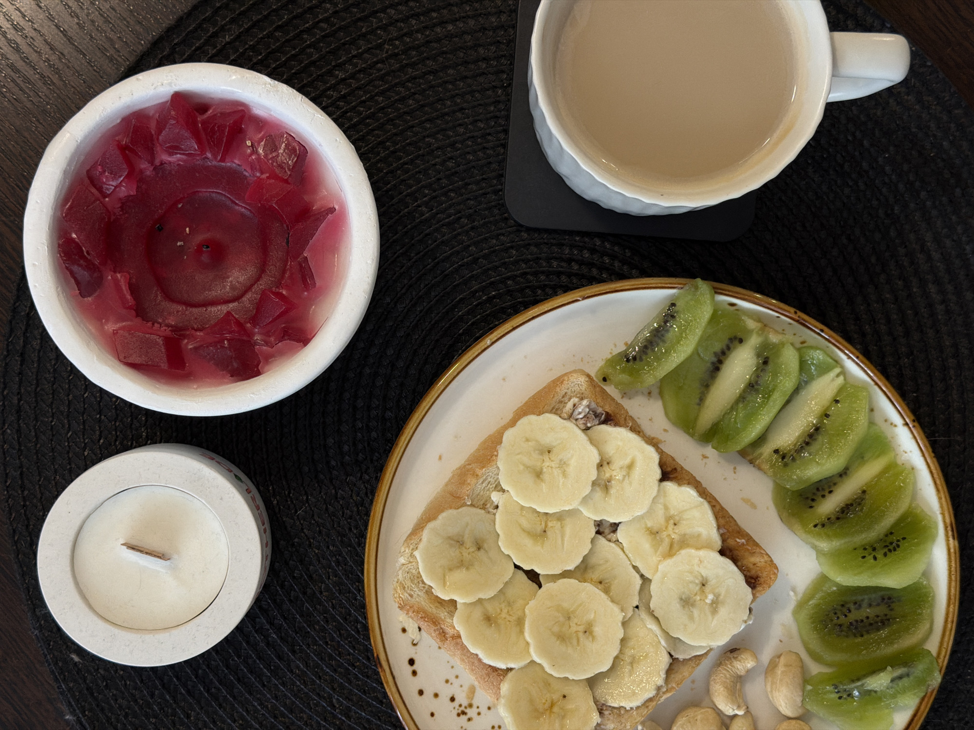 This overhead shot captures a cozy and healthy meal arrangement laid out on a dark, possibly wooden, table, set against a black woven placemat. The scene is located in Засечное, Russia.
In the upper left, a white ceramic bowl holds a deep red, melted candle with irregular chunks of solidified wax around its edges. Below it, a smaller, light-colored circular holder contains an unlit candle with a distinct wooden wick. Faint red text, possibly "MERRY", is visible on the side of this holder. To the upper right, a white ceramic mug, placed on a black square coaster, contains a light beige beverage, likely coffee or tea with milk.
The main subject of the image is a white ceramic plate with a brown rim, positioned in the lower right. It holds a slice of lightly toasted bread topped with sliced bananas, suggesting a spread underneath. Alongside the toast, numerous slices of green kiwi fruit are neatly arranged in a curved line. A small pile of cashew nuts sits at the bottom of the plate, adding to the nutritious selection.
The overall impression is one of a wholesome and peaceful breakfast or snack, presented attractively with natural daylight illuminating the scene. The items suggest a relaxed moment, possibly a self-care routine.