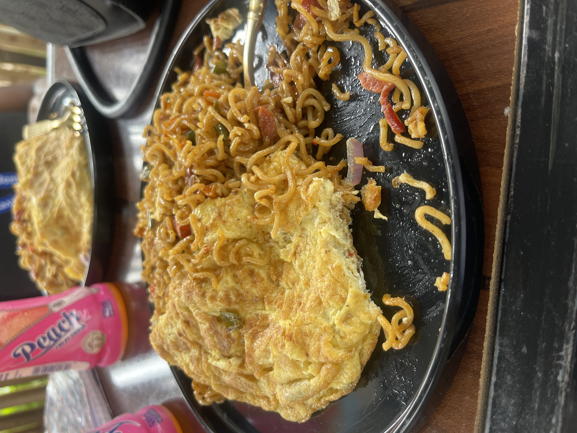 A plate of food is displayed on a wooden table. The plate contains noodles mixed with vegetables and what appears to be an omelet. The noodles are thin and curly, with visible pieces of red pepper, green pepper, and red sausage. The omelet is golden brown and folded over a portion of the noodles. The plate is black and slightly worn. In the background, another plate with food is partially visible, as are two pink bottles with text. One bottle clearly shows the word "Peach" in a stylized font. The scene appears to be outdoors, suggested by the blurred greenery in the background and the rustic wooden table. The lighting is natural, suggesting it is daytime. The food is the central focus of the image, presented in a close-up view. The overall impression is of a casual meal, possibly in a relaxed outdoor setting.