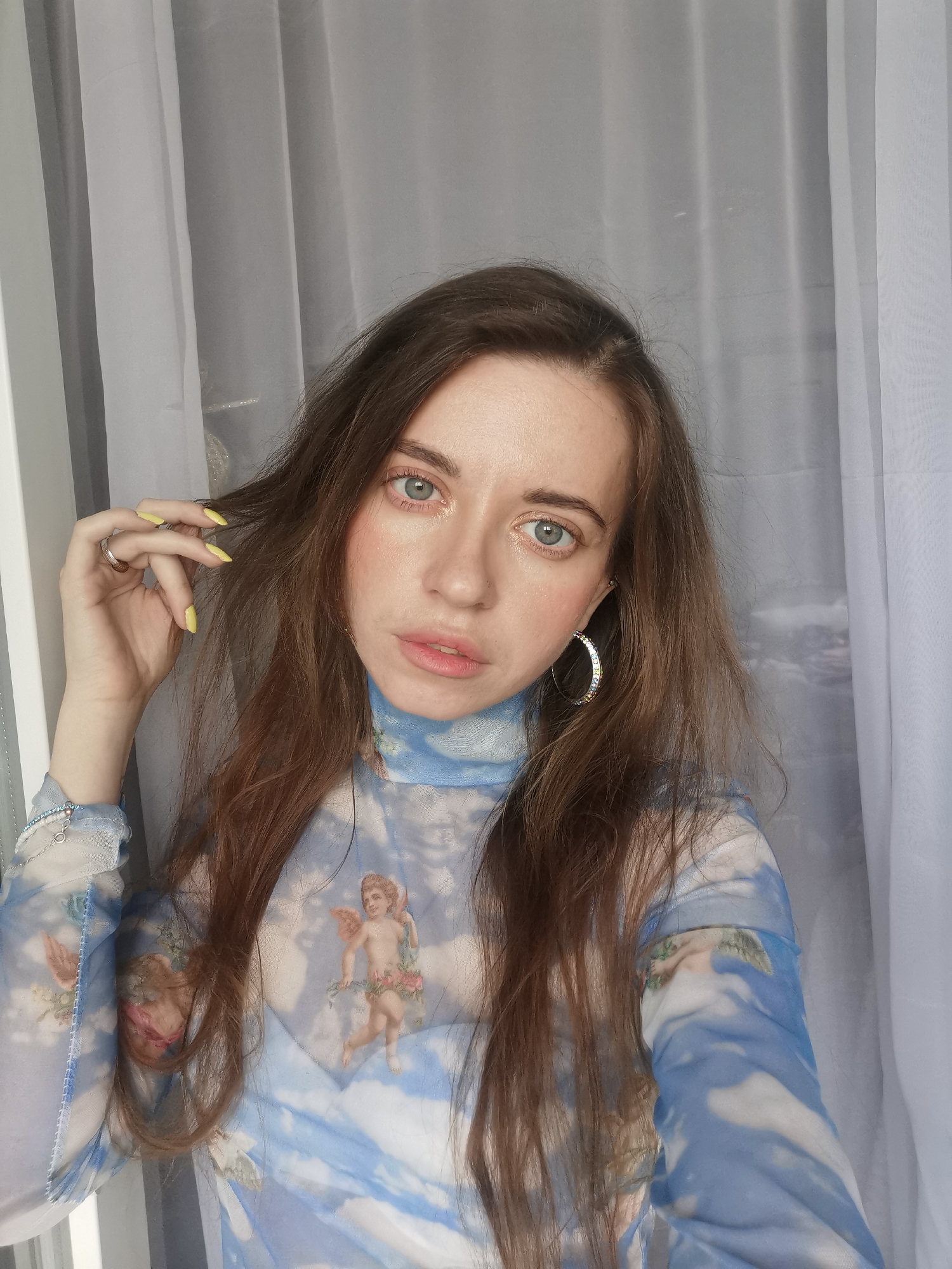 A light-skinned woman with long brown hair is centered in a portrait-style shot. She wears a semi-sheer top printed with a blue sky and cherub motifs, and a large silver hoop earring is visible on her left ear. Her left hand, adorned with a silver ring and yellow nail polish, is raised near her hair. Behind her are sheer, white-gray curtains. The location is indoors in an unknown city and an unknown country.