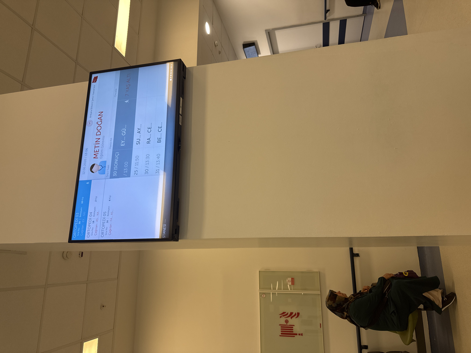 The image shows a digital display screen in a hospital waiting area. The screen displays patient information including appointment numbers, names, and estimated wait times. It indicates that the location is Ankara Bilkent Şehir Hastanesi, which is a city hospital in Ankara, Turkey. The display also shows the date and time as 12.06.2026 / 13:06. Below the main display, there is a smaller screen or label that says "STANESİ". To the left of the display screen, a woman wearing a headscarf and a dark green robe is sitting on a green chair, looking to her left. Behind her, on the wall, is a fire safety sign depicting a coiled fire hose and instructions. The environment appears to be an indoor hallway or waiting room with tiled floors and white walls. A fire alarm detector is visible on the ceiling.