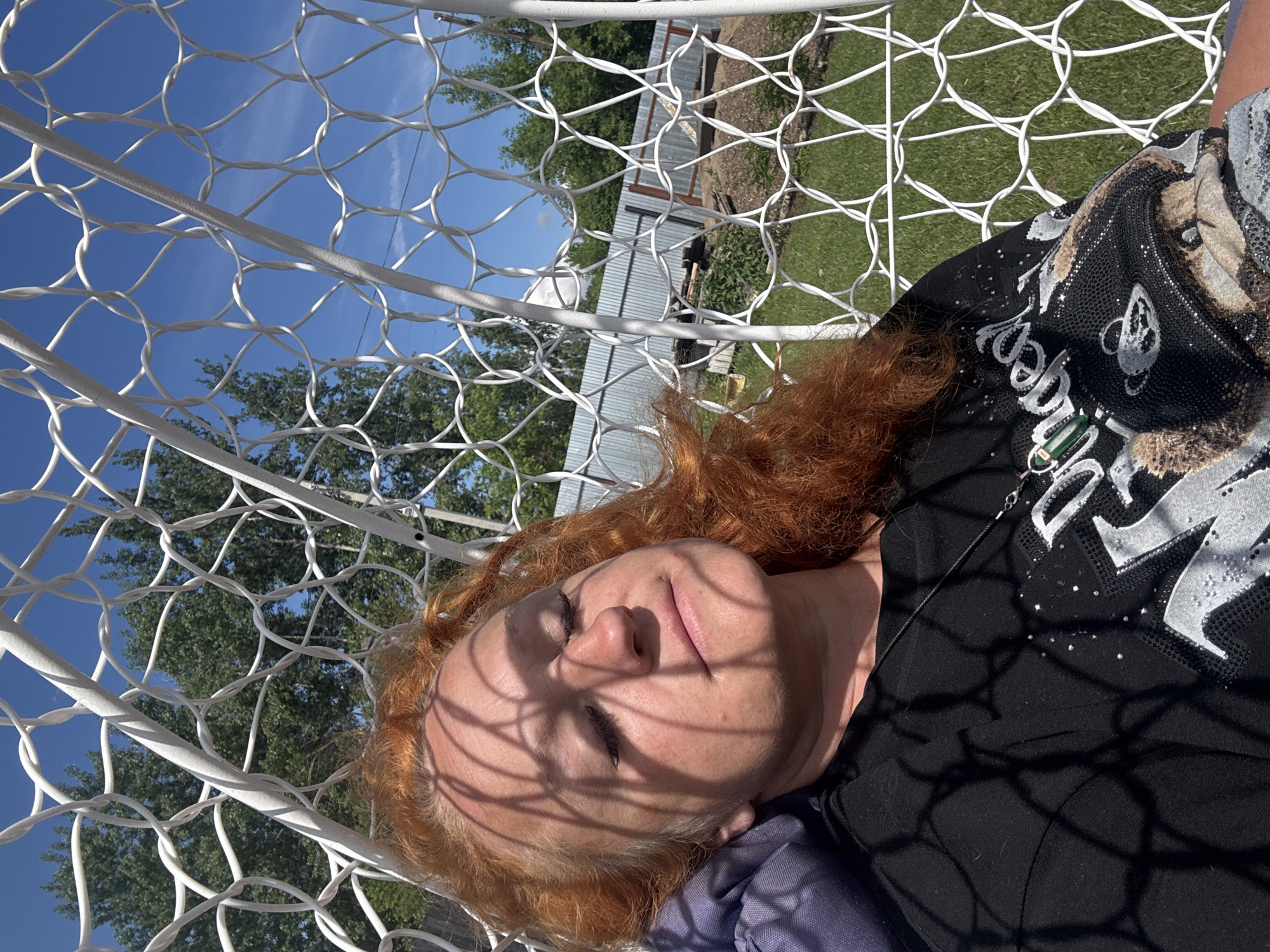 An eye-level outdoor photograph captures a person, likely an adult female, with distinctive reddish-orange, curly hair, resting in a white net hammock or hanging chair. Her eyes are closed, conveying a sense of peace and relaxation, and pronounced, geometric shadows from the net fall across her face and body. The bright, clear blue sky and strong shadows indicate a sunny midday or afternoon. She wears a black t-shirt embellished with stylized, glittery text and graphic elements, which appear to spell "Disney". The background features vibrant green trees and grass, and a distant corrugated metal outbuilding. This tranquil scene depicts a moment of outdoor leisure, possibly in a garden or backyard in Chaikovsky, Russia.