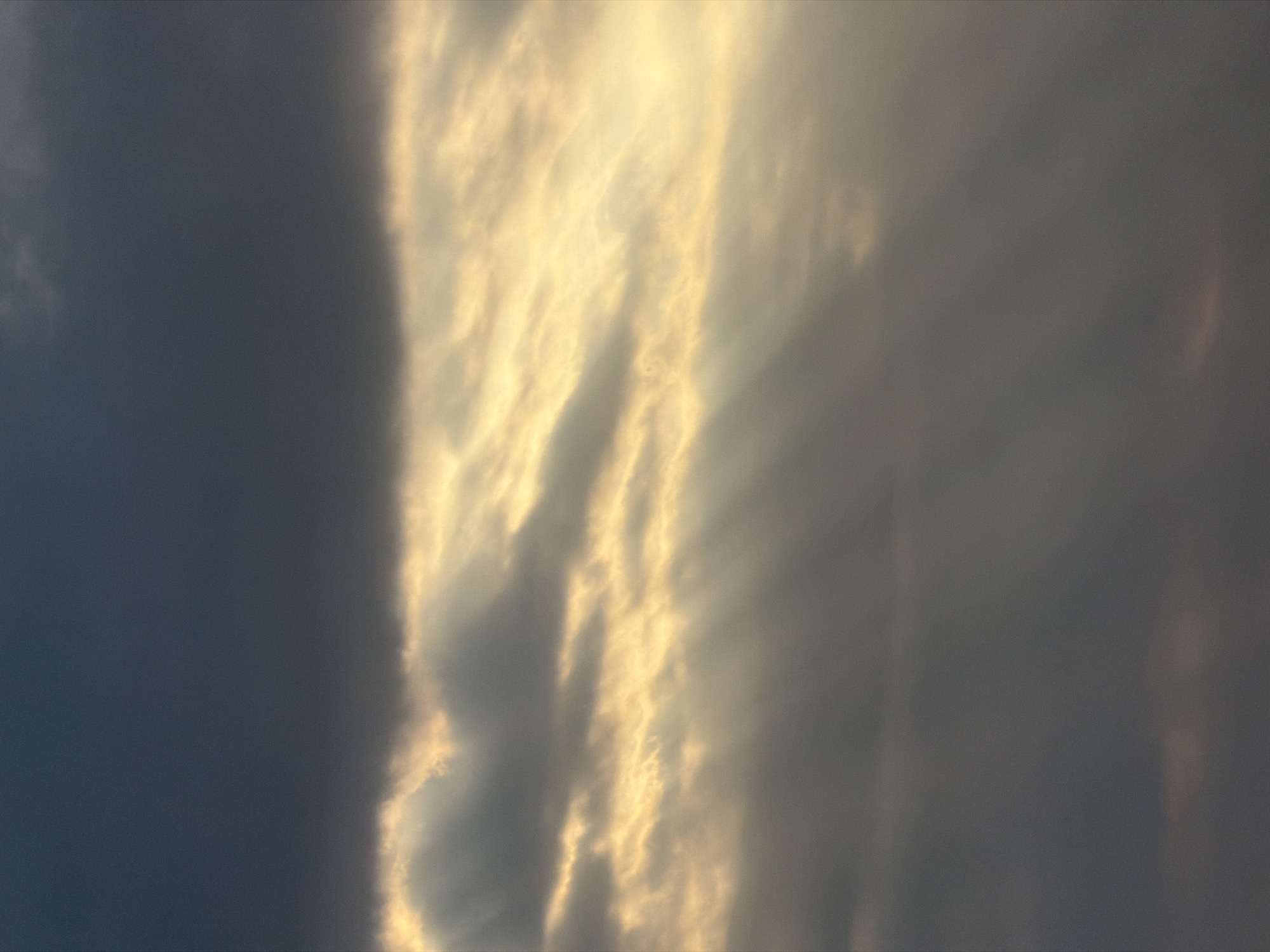 The image displays a dramatic sky scene, primarily composed of dense clouds and striking sunlight. On the left, a significant portion of the frame is filled with dark, heavy clouds, creating a stark contrast with the vibrant, golden light that slices vertically through the center. This light appears as intense, radiant shafts, characteristic of crepuscular rays, illuminating the intricate textures and edges of the clouds with a brilliant yellow-orange hue.
Further to the right, the light diffuses into more subtle rays, blending into a lighter, grayish cloud cover. The overall impression is one of a powerful natural phenomenon, likely captured during either sunrise or sunset due to the rich, warm color palette of the light against the darker clouds. There are no people, man-made objects, or visible text within the frame. The scene evokes a sense of awe and the majestic beauty of light breaking through cloud cover, located in North Kuta, Indonesia.