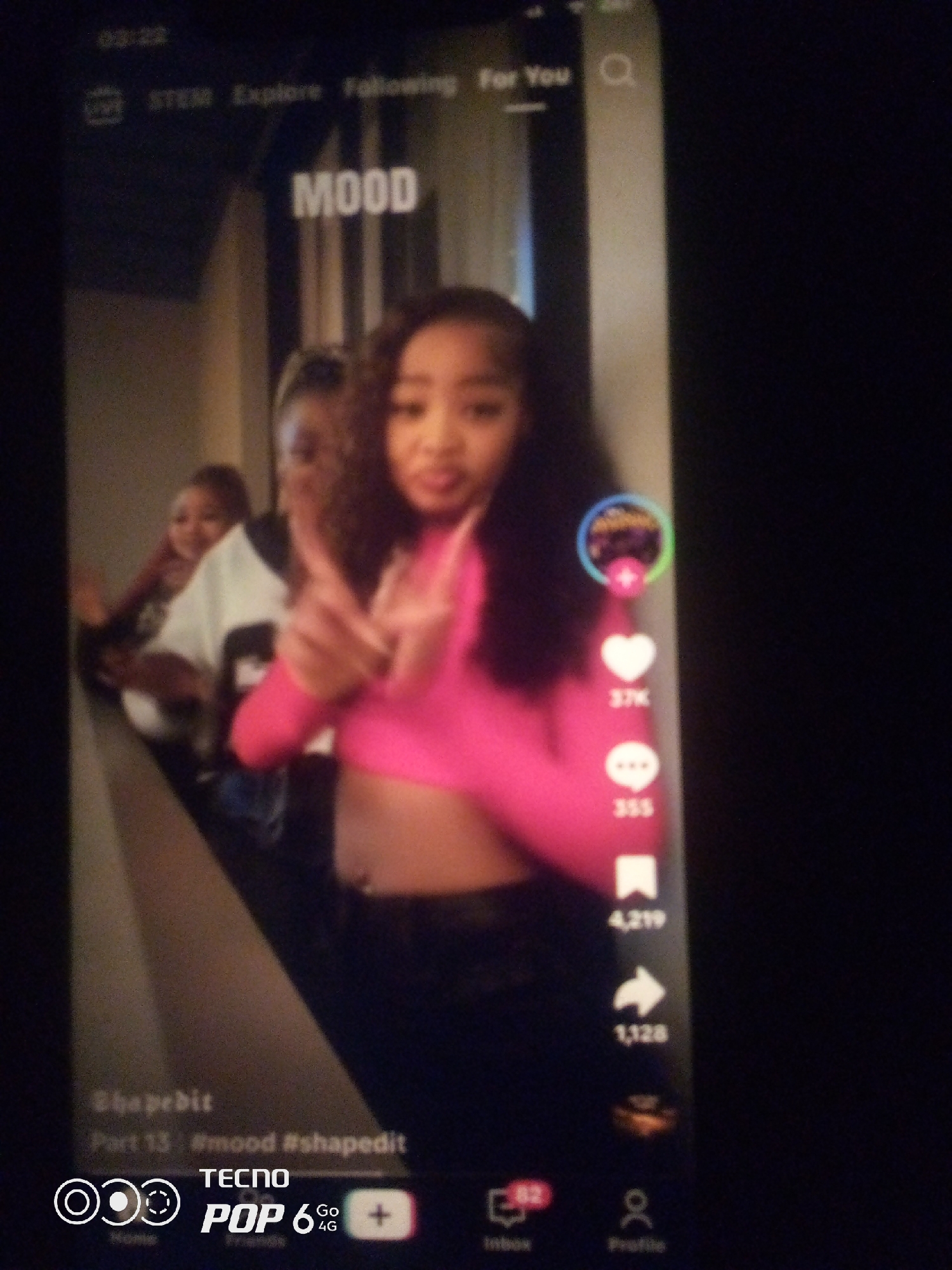 The image is a screenshot of a TikTok video showing three young Black individuals. The person in the foreground is a female wearing a bright pink top, looking at the camera with her hands crossed in a "V" shape. Behind her, two other individuals are visible, one wearing a white shirt and the other with darker clothing. The video appears to be a dance or lip-sync video, as indicated by the text "MOOD" at the top of the screen and the engagement icons on the right side, including likes, comments, and shares. The bottom of the screenshot shows the user interface of the TikTok app, with options for "Home," "Inbox," and "Profile." The bottom edge of the phone also displays "TECNO POP 6 Go 4G." The lighting suggests an indoor setting. The image quality is low and blurry. The location is not specified in the image.