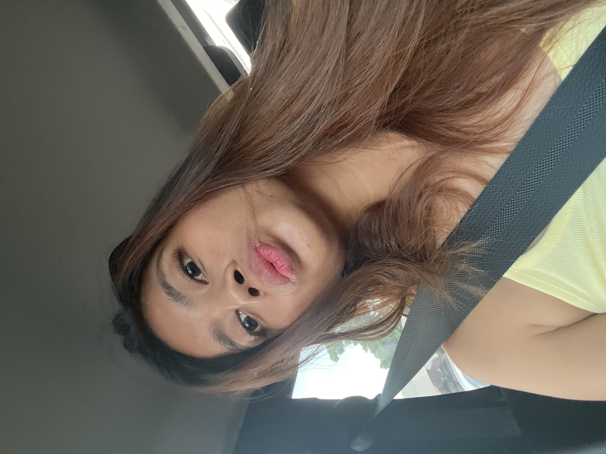 A young woman with long brown hair is seen in the passenger seat of a car, wearing a yellow tank top and a black seatbelt. She is leaning towards the camera, with her lips pursed in a kiss pose. Her eyebrows are dark and arched, and she has on what appears to be subtle eye makeup. The background suggests an outdoor environment, possibly a street or residential area, with trees visible through the car window. The lighting is natural, indicating it's daytime. The image appears to be a selfie taken inside a vehicle in Davao City, Philippines.
