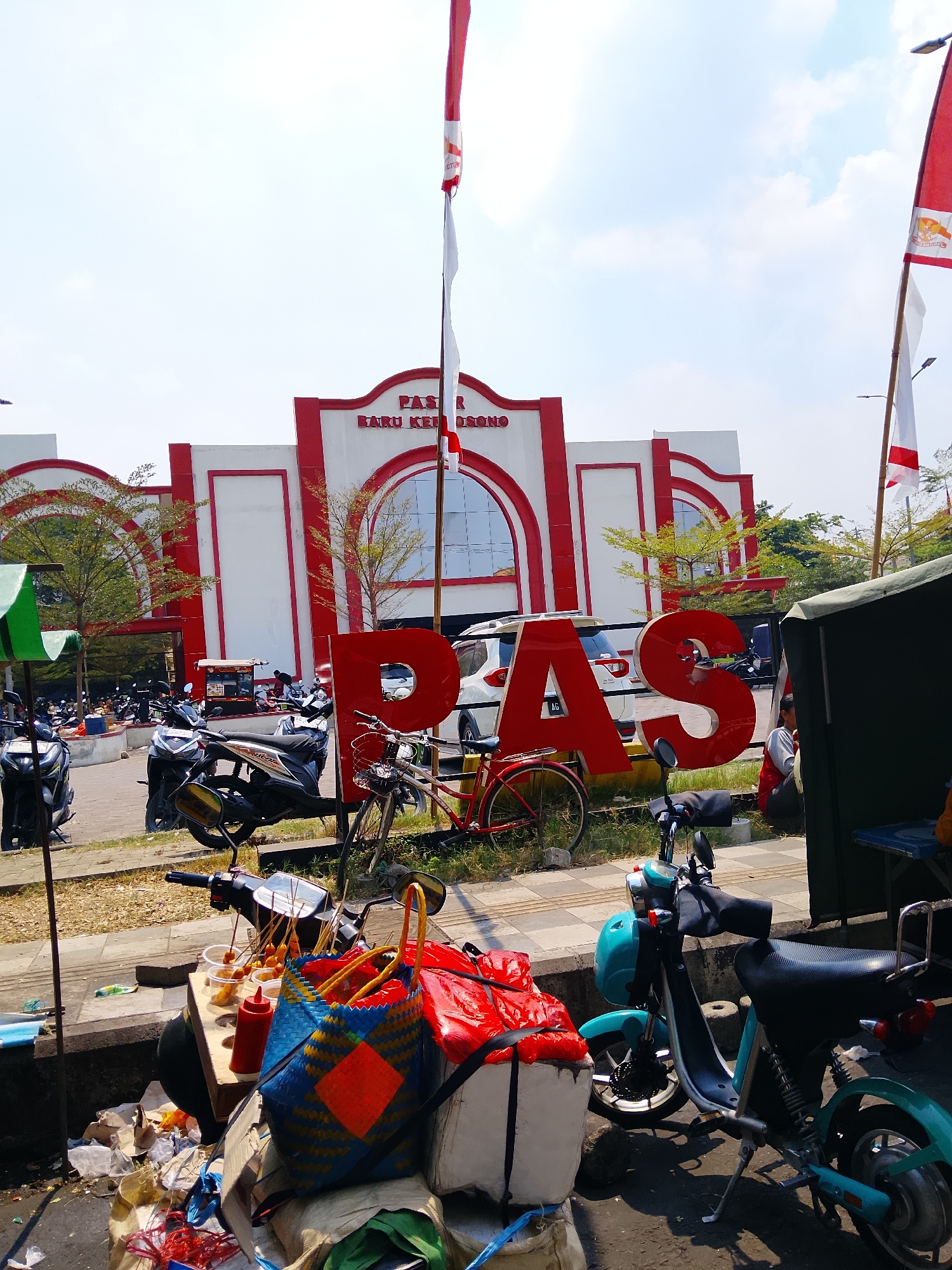 The image depicts the entrance to a market in Kertosono, Indonesia, identified by a large red sign spelling "PAS" in front of the building. The building itself is white with red trim, featuring arched windows. Indonesian flags fly from poles in front of the building. In the foreground, there are several parked motorcycles, a red bicycle, and a food stall displaying snacks on skewers, sauces, and bags. A turquoise electric scooter is also parked in the foreground. The scene appears to be outdoors on a sunny day, with some greenery and scattered debris visible.