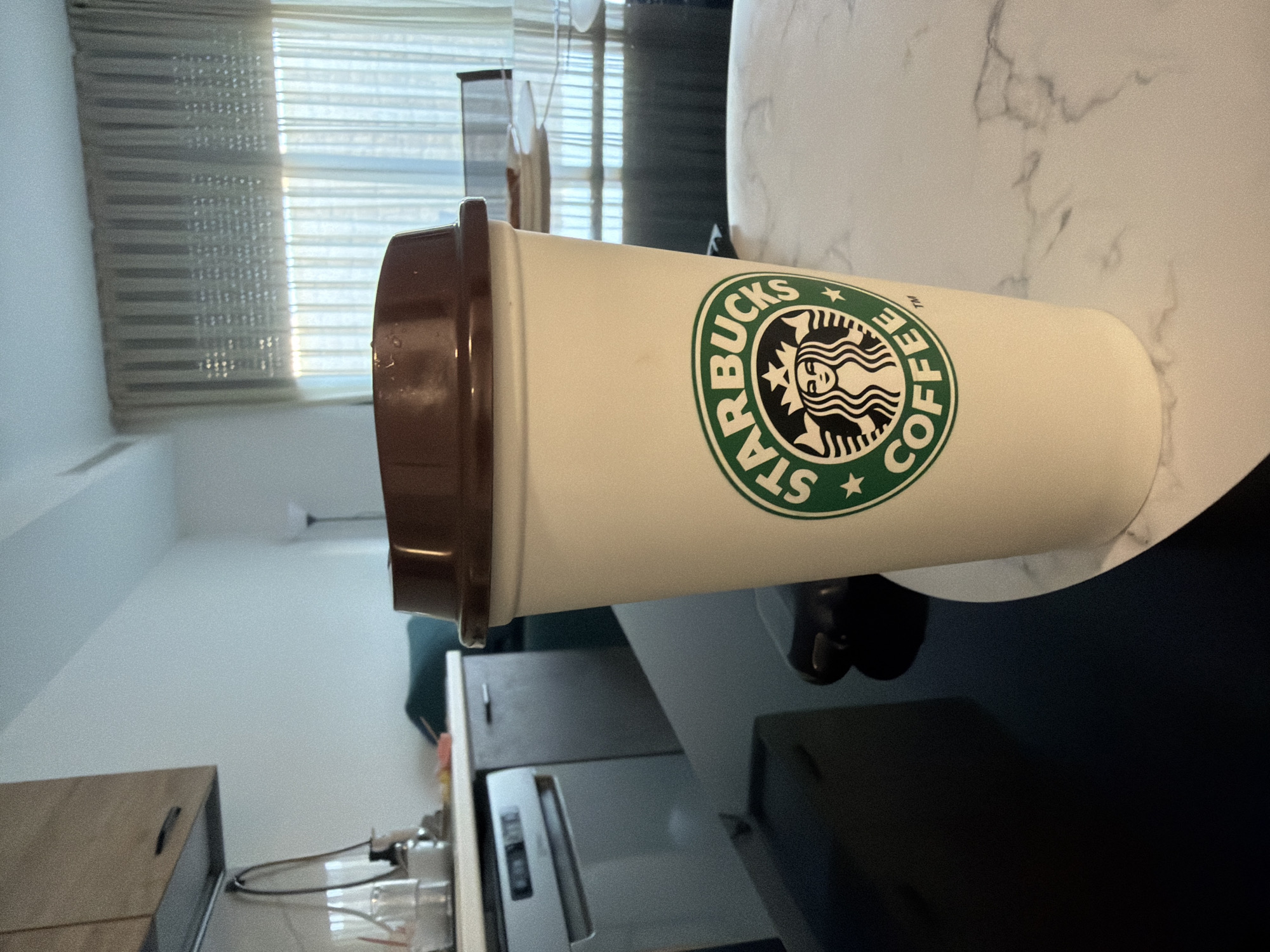 The image shows a Starbucks coffee cup on a marble-patterned table. In the background, a kitchen counter with cabinets, a sink, and a dishwasher is visible. Further back, sheer curtains cover a window, and a floor lamp stands in the corner. The scene appears to be in a residential apartment or hotel room, possibly located in Finestrat, Spain, given the context of the location being Finestrat, Spain. There are no people visible in the image.