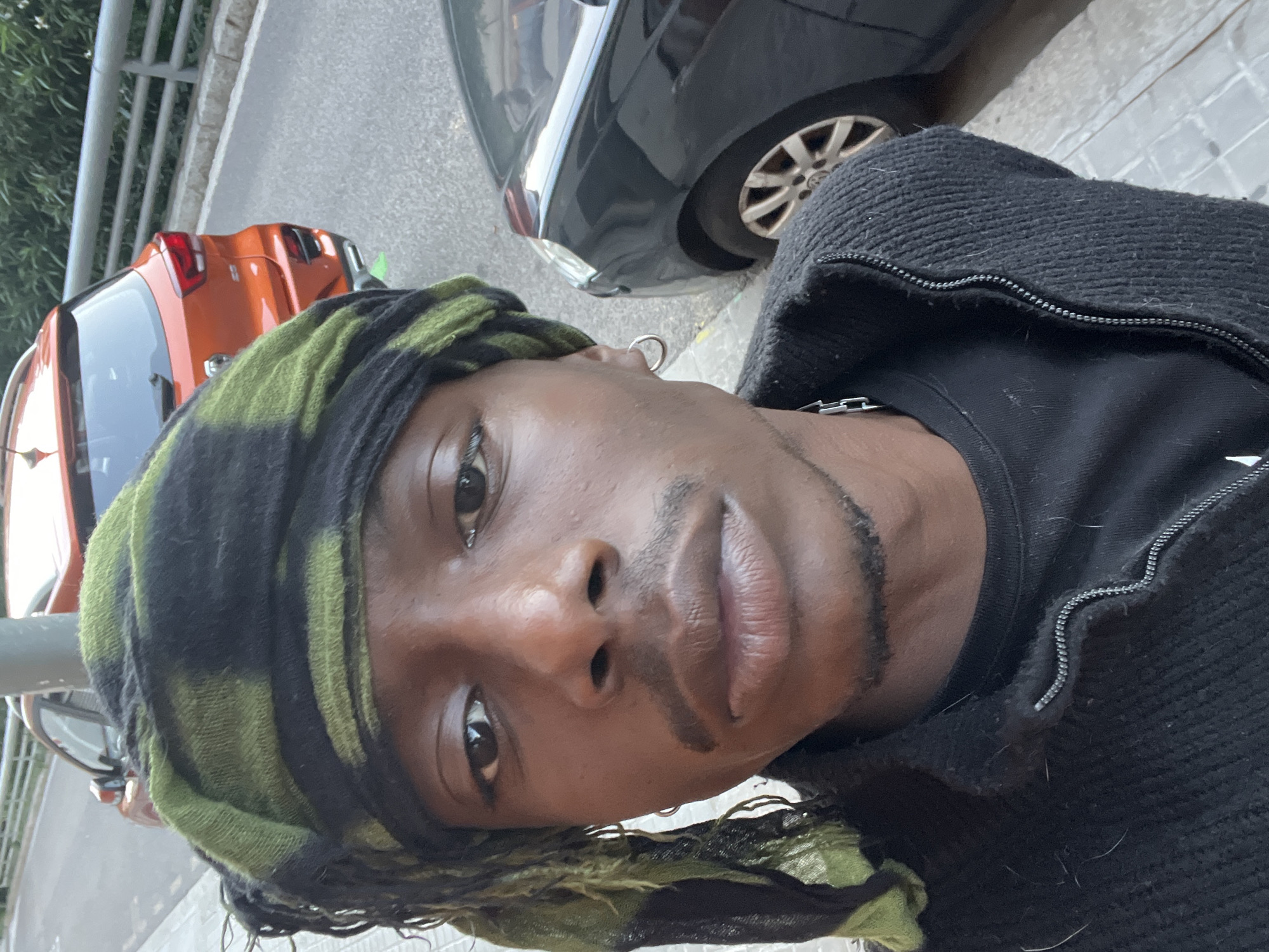 A close-up, ground-level perspective photo shows a person of color, likely male, wearing a green and black headscarf, a black T-shirt, and a black zippered sweater. The person has a silver earring. The background includes the street scene with parts of two cars, one orange and one dark gray/black. The setting appears to be Barcelona, Spain, given the architectural style and vegetation. The photo is taken at an angle.