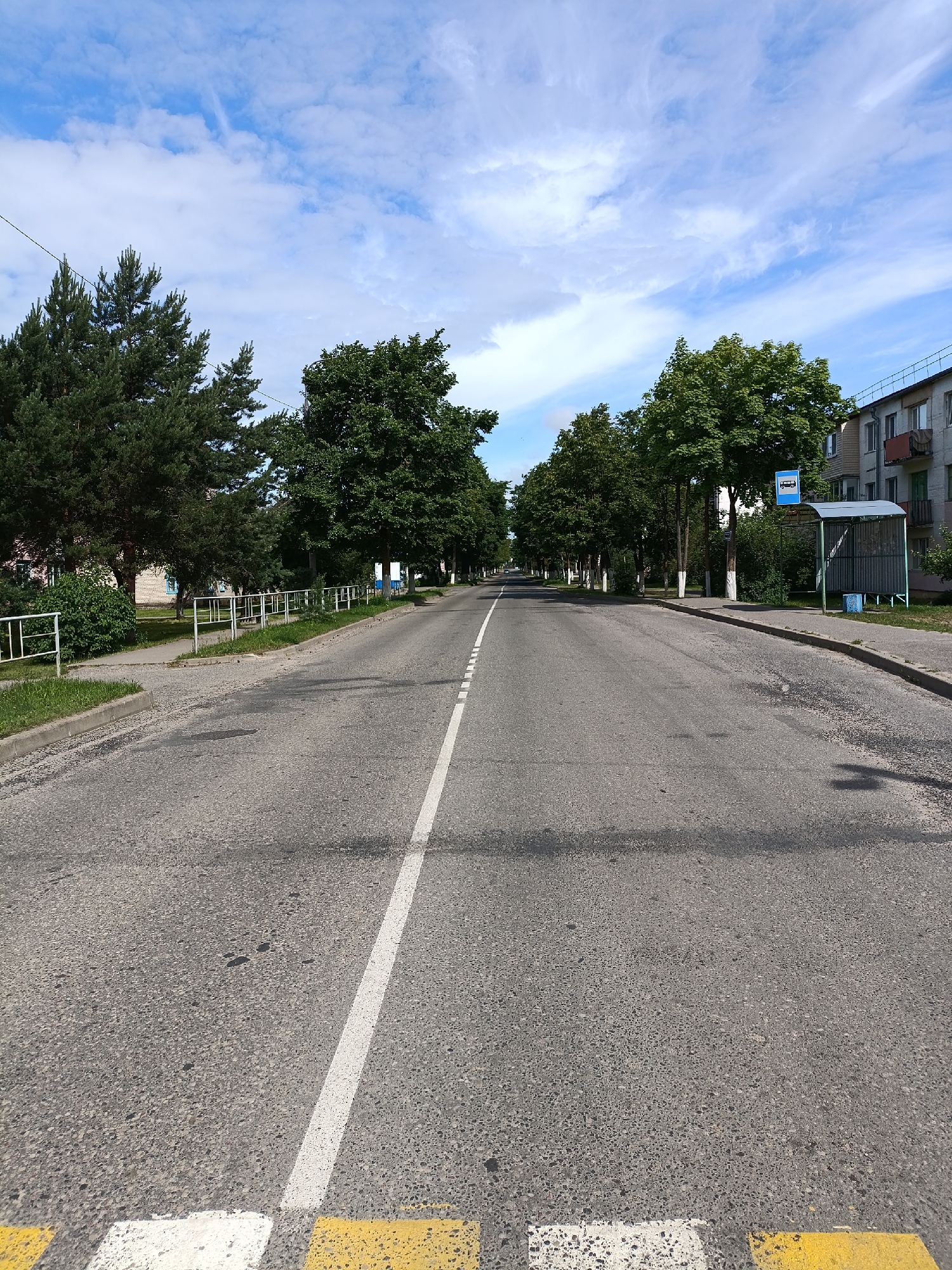 The image depicts a wide, paved road in a town, lined with trees on both sides. The road has a white lane marker down the center and appears to be a straight, flat stretch. On the left side of the road, there is a sidewalk with a low white fence and some greenery. On the right side of the road, there is a bus stop with a shelter and a blue bus stop sign. Further down the road on the right, a multi-story building with visible windows and balconies is present. The sky above is blue with scattered white clouds. The foreground shows a portion of the road with a pedestrian crossing marked by yellow and white stripes. There are no people visible in the image.