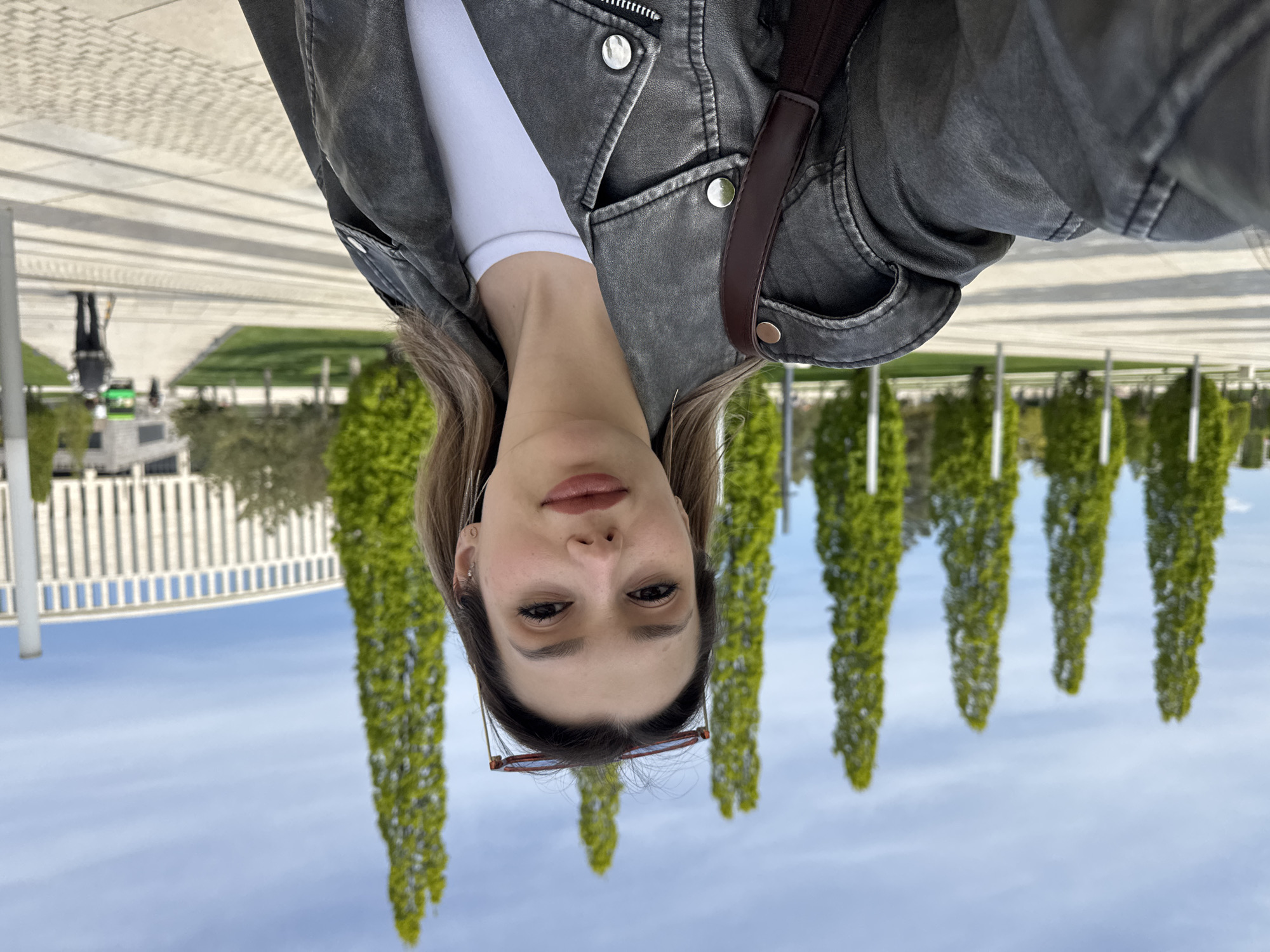 An upside-down selfie captures a young woman in the foreground, looking directly at the camera. Her face and upper body are prominent, featuring light brown hair and a neutral expression. She wears a dark grey, textured jacket over a white long-sleeved top, with a dark brown strap (likely from a bag) visible on her right side. Red-framed glasses are perched on her head.
The background, also inverted, depicts an outdoor public space under a clear, light blue sky. A prominent row of tall, slender green trees with dense foliage stretches across the middle ground. Beyond the trees, modern architecture is visible, including white buildings with vertical slatted designs. Paved areas with geometric patterns and some green spaces suggest a well-maintained park or urban plaza. Another person is visible in the distant background, also inverted. The scene is brightly lit, indicating daytime. There is no visible text.