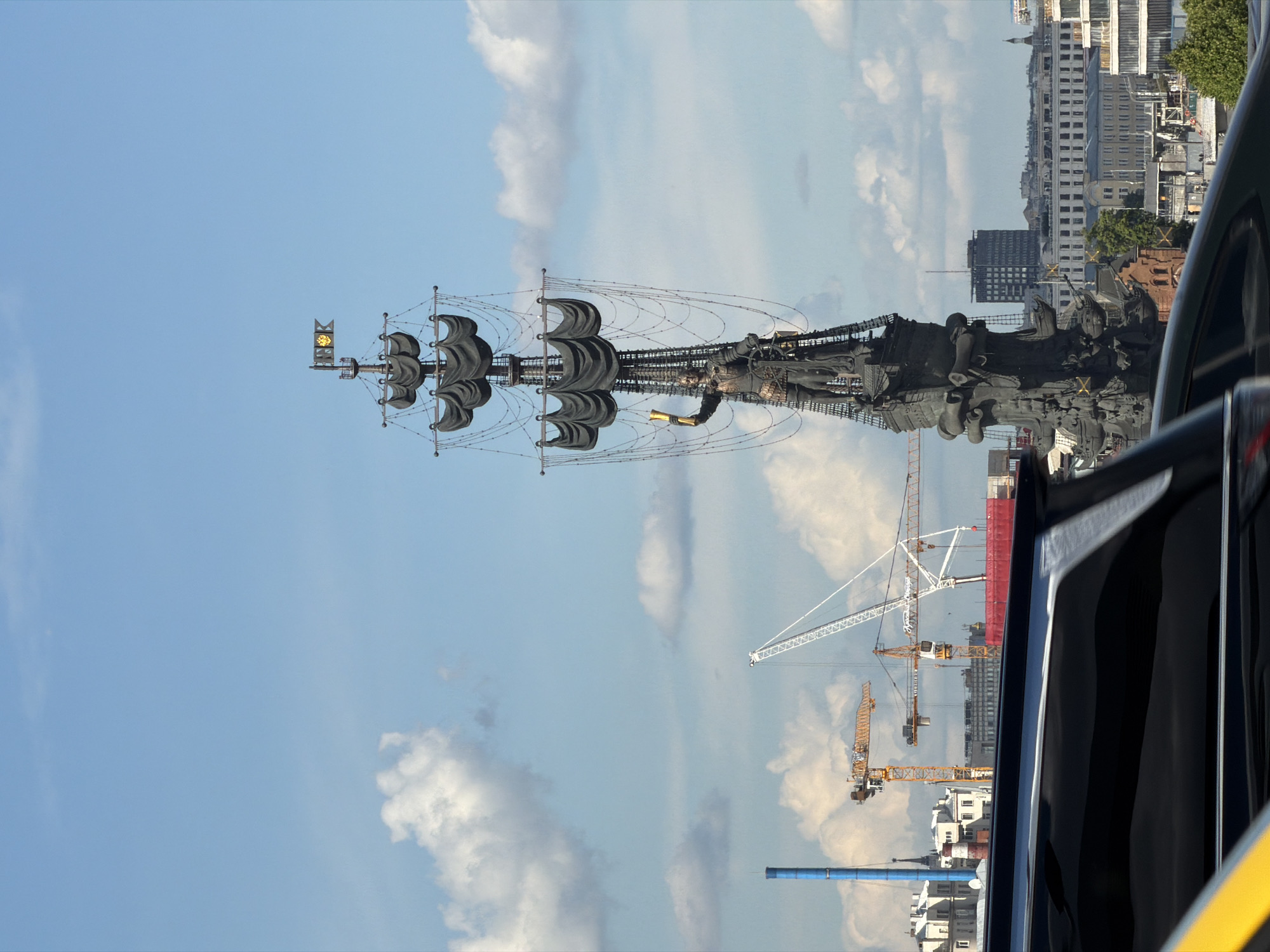 The image features the monumental Peter the Great Statue in Moscow, Russia, set against a bright, partly cloudy sky. The impressive bronze statue depicts Peter the Great standing on the deck of a large sailing ship, complete with three masts, numerous furled sails, and intricate rigging. Peter the Great is shown in imperial military attire, holding a spyglass in his right hand. A distinctive golden emblem, resembling a sun or compass rose, is prominently displayed on the mainmast of the ship. The base of the monument also features smaller figures, likely sailors or allegorical representations.
In the background, a sprawling cityscape is visible, characterized by a mix of buildings and active construction sites. Several large cranes, including both white and yellow variants, punctuate the skyline, indicating ongoing urban development. A tall, slender blue cylindrical structure is also visible among the buildings. The photograph captures the scene during daytime, with clear visibility and ample sunlight illuminating the statue and the surrounding environment. White, fluffy clouds are scattered across a wide expanse of blue sky.
The perspective of the shot suggests it was taken from a vehicle, with the dark frame of a car door or window and a yellow stripe visible in the bottom right corner of the image. The scene presents a striking juxtaposition of historical grandeur with contemporary urban expansion. No discernible text is visible within the image.