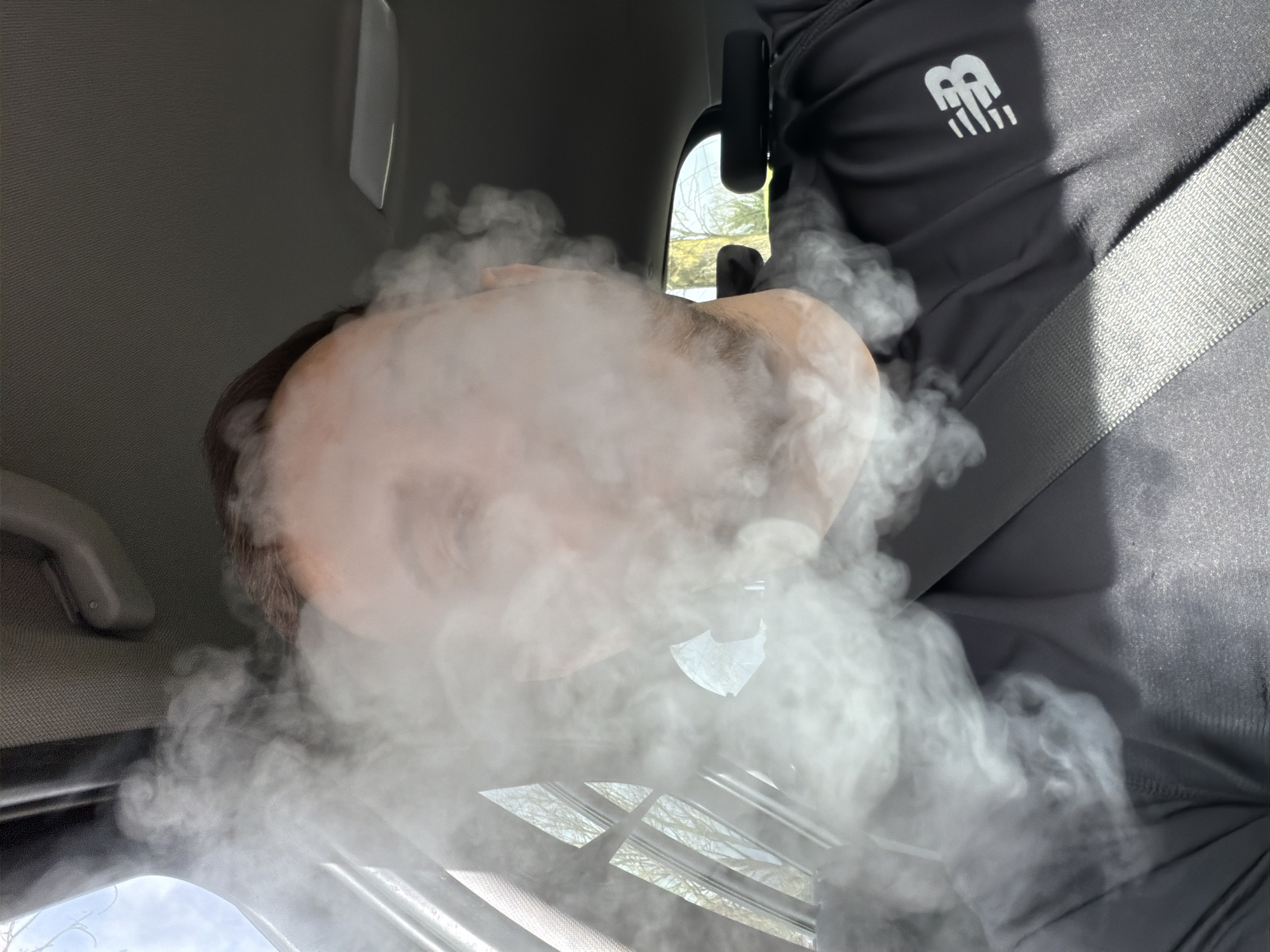 A person is exhaling a large cloud of vapor while seated in a vehicle. The person is wearing a dark grey athletic top with a white logo on the right shoulder and a grey seatbelt across their chest. The vehicle interior is grey. The vapor obscures much of the person's face. The vehicle's sun roof and interior lights are visible. The surroundings outside the vehicle are blurry, suggesting motion. There is no evidence to suggest this photograph was taken in London.