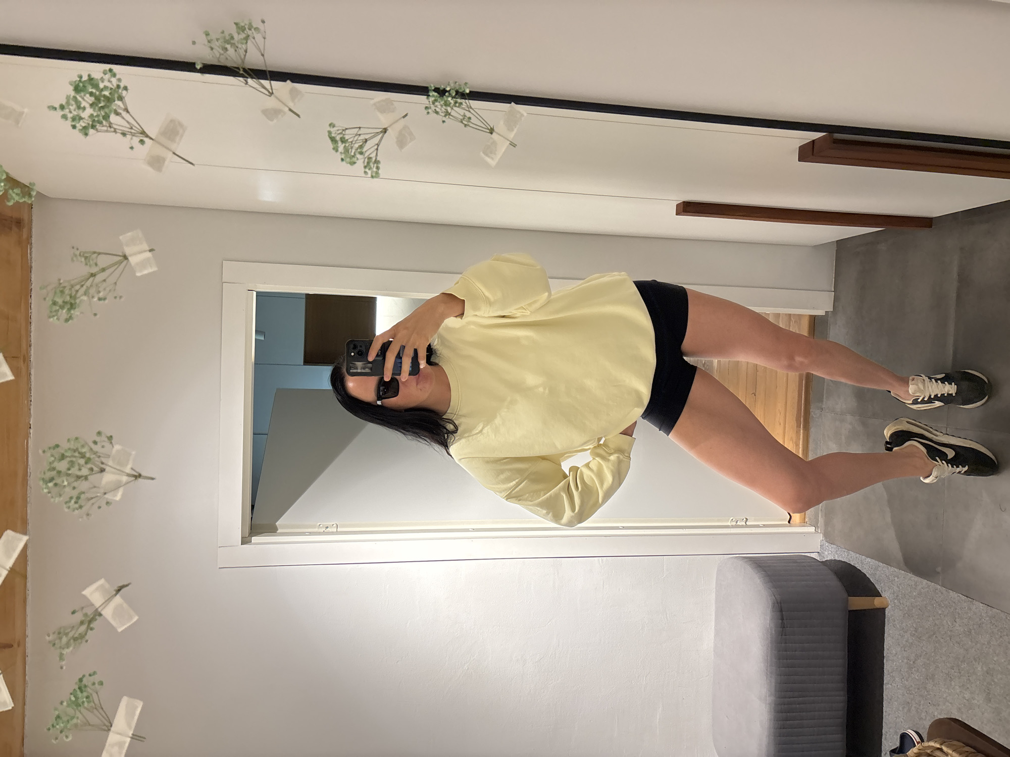 A woman is taking a mirror selfie indoors. She is wearing a light yellow long-sleeved shirt and black shorts. Her hair is black and long. She is holding a black smartphone in her left hand, presumably to capture the photo. The reflection shows a room interior with a doorway, a stool, and decorations of taped baby's breath on the mirror and walls. The floor is gray. The location and city are not discernible.