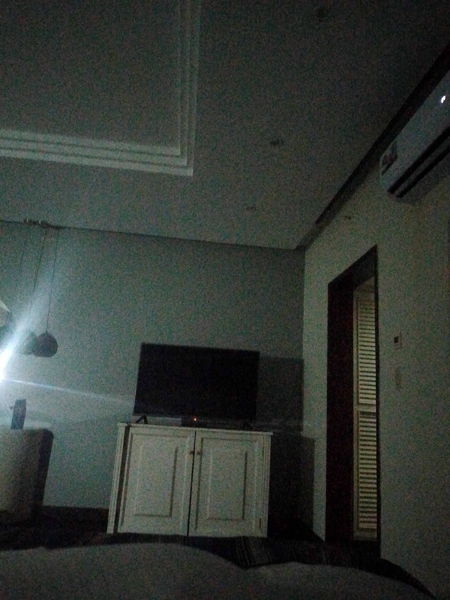 The image shows an interior shot of a room, likely a living room. It appears to be nighttime or a dimly lit space. The room contains a television set placed on top of a white cabinet. There is a doorway visible to the side of the television, with a window/shutter seen through it. In the foreground, there is furniture which appears to be a bed or large sofa. The ceiling has some design details including what appears to be a trim/border. An air conditioning unit can be seen mounted on the upper right wall. Pedro González, Venezuela cannot be determined from the image.