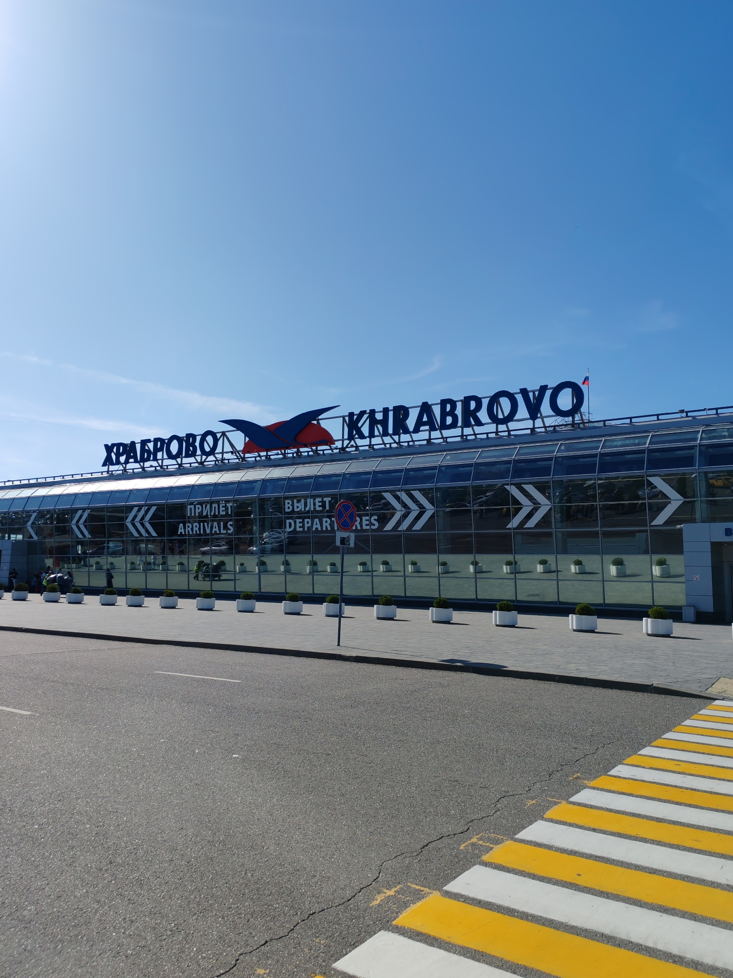 The image shows the exterior of Khrabrovo Airport in Russia on a clear, sunny day. The building features a modern glass facade with large windows reflecting the sky and surrounding environment. Above the glass, large blue letters spell out "KHRABROVO," with a stylized red and blue bird logo integrated into the signage.
Visible text on the building's glass includes "ПРИЛЁТ ARRIVALS" and "ВЫЛЕТ DEPARTURES." The building is lined with white planters containing small green bushes, and a paved walkway runs in front of it. To the right of the building, a blue and red circular sign with a camera symbol indicates a no-photography zone.
In the foreground, a section of asphalt road is visible, leading to a brightly marked yellow and white pedestrian crossing. A few people are seen on the walkway in front of the airport, some appearing to be with luggage, suggesting travelers are arriving or departing. The overall atmosphere is bright and open, typical of an airport setting on a pleasant day. The Russian flag is visible flying on a flagpole atop the building.