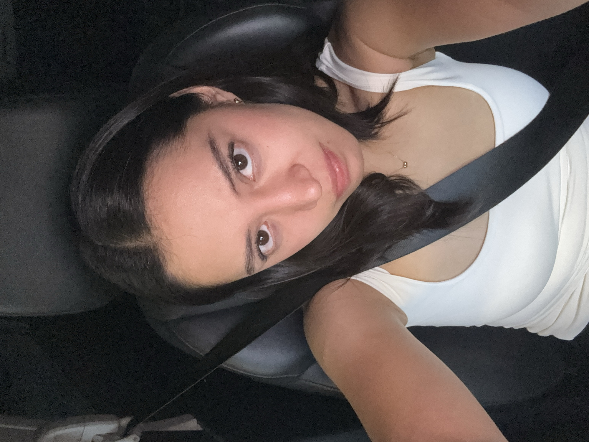 A woman with dark hair and brown eyes is sitting in a car. She is wearing a white top and a seatbelt is fastened across her chest. The car's interior is dark, with black leather seats. The location is unknown and cannot be determined from the image.