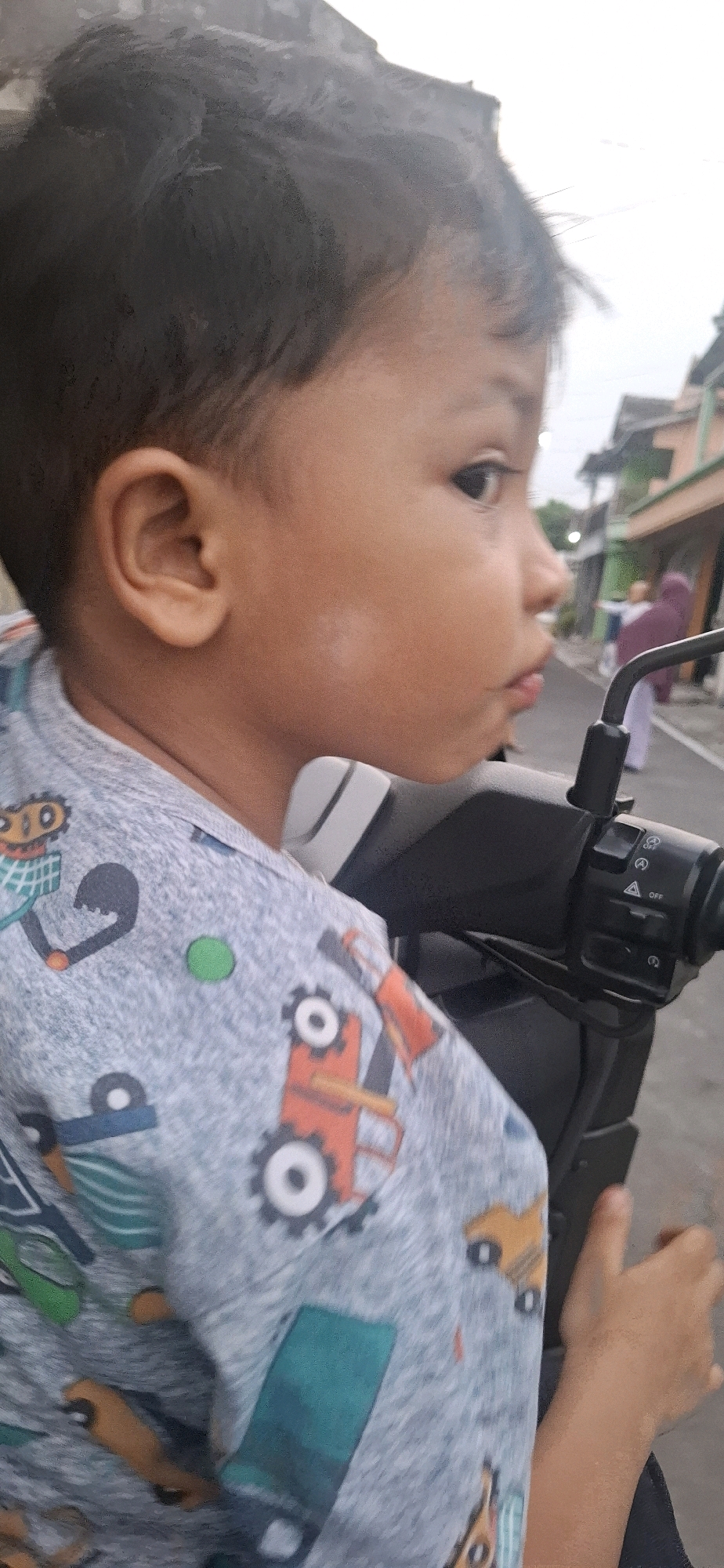 A young child is captured in a close-up, side-profile shot, seemingly riding on a motorcycle in Desa Gagaksipat, Indonesia. The child's face, with brown skin and dark hair, is turned to the right, with eyes looking slightly upwards. A light-colored patch is visible on the child's left cheek. The child is wearing a light gray shirt adorned with a pattern of colorful toy vehicles, including orange and yellow trucks and blue and green construction equipment.
The child's left hand is resting on the handlebars of the motorcycle, which are visible in the foreground and are black and gray. The handlebars have various controls and labels, including "OFF" and what appear to be symbols for lights or indicators. The background suggests an outdoor setting, possibly a street or road, with houses and people in the distance. One person in the background is wearing a purple headscarf. The lighting indicates it is daytime, possibly late afternoon or early morning due to the softness of the light and slightly overcast sky. The overall impression is a casual moment of a child enjoying a ride, perhaps with a family member, in a residential area.