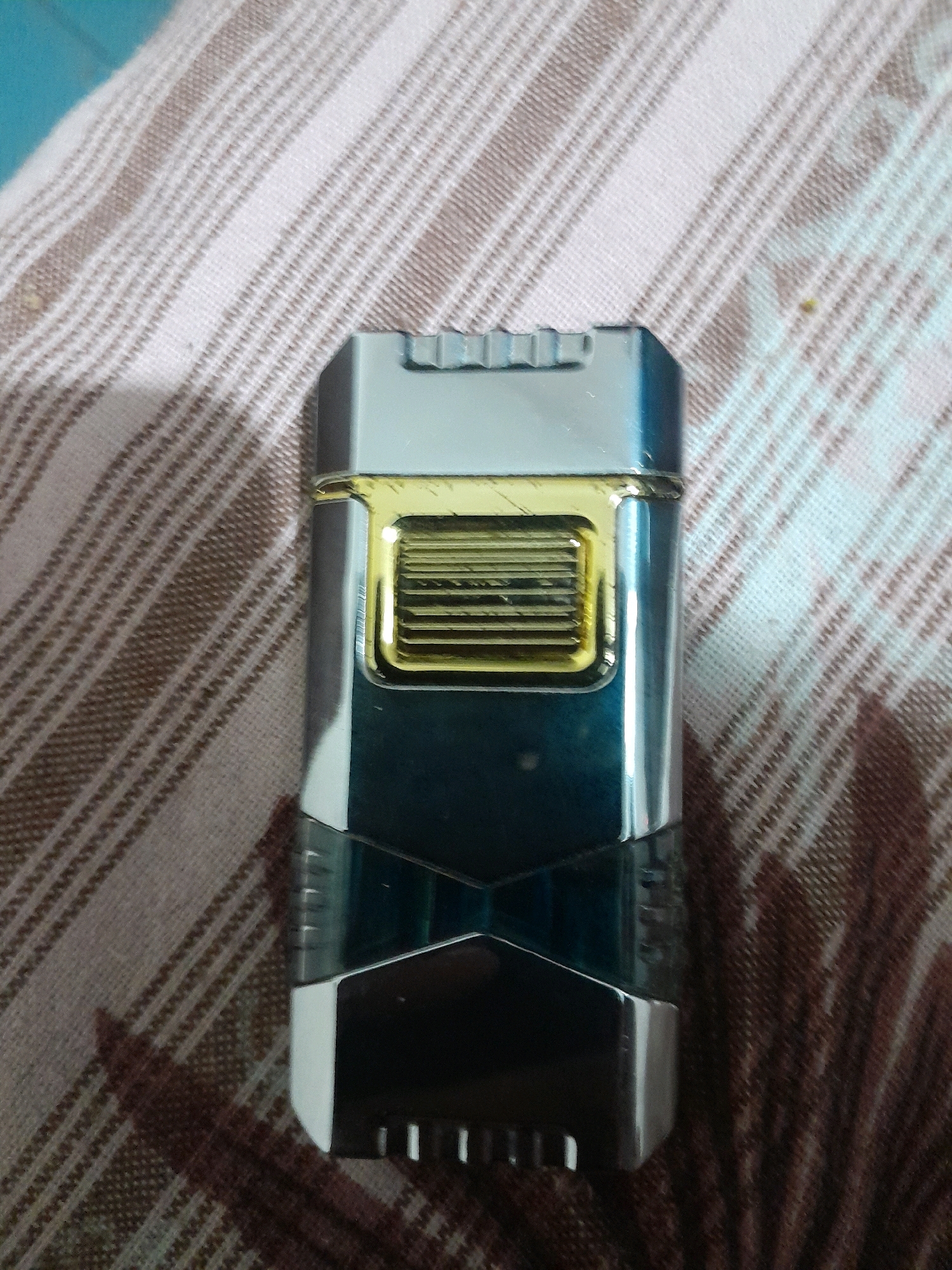 A silver and blue electric shaver with a gold grill is lying on a beige and white striped fabric. The shaver is oriented vertically, with its top facing upwards. The fabric appears to be a tablecloth or a blanket. The scene is indoors, as indicated by the artificial lighting. No people are visible in the image. The specific location in Kadalundi, India, is not discernible from the visual information provided.