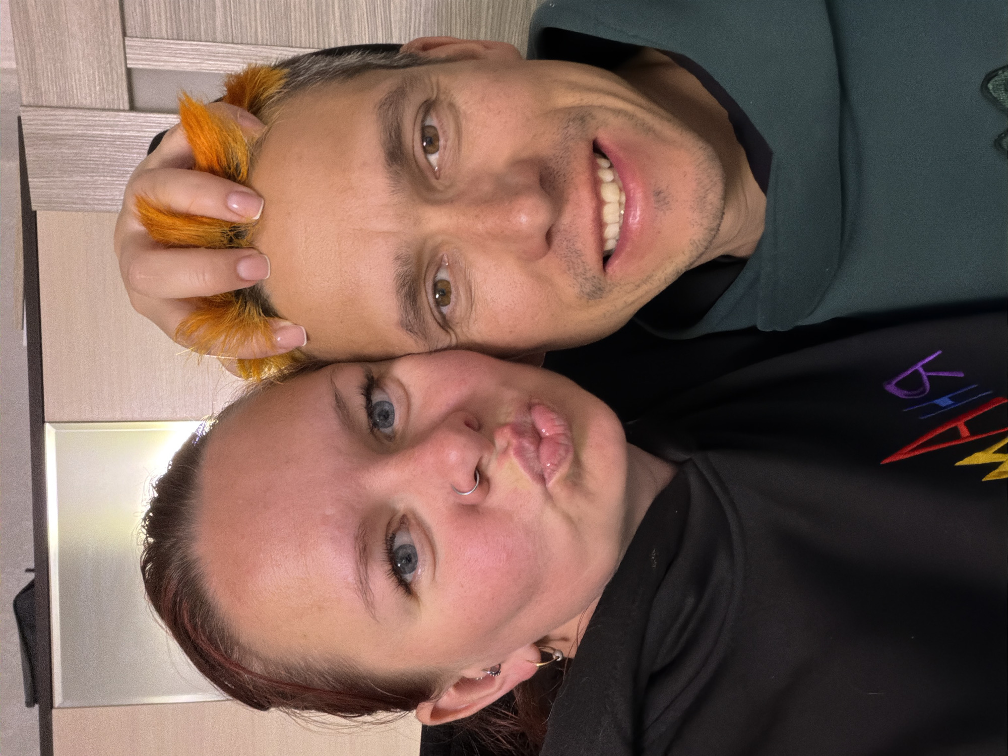 Two people are posing closely for a photo. A man with light skin and short, bleached hair that is dyed orange at the front is smiling and looks directly at the camera. He is wearing a dark green hoodie. A woman with fair skin, light blue eyes, a nose ring, and pursed lips is positioned below the man. She is wearing a black top with some colorful embroidery on the right side. They appear to be indoors. No specific details indicate the city of Chaikovsky, Russia.
