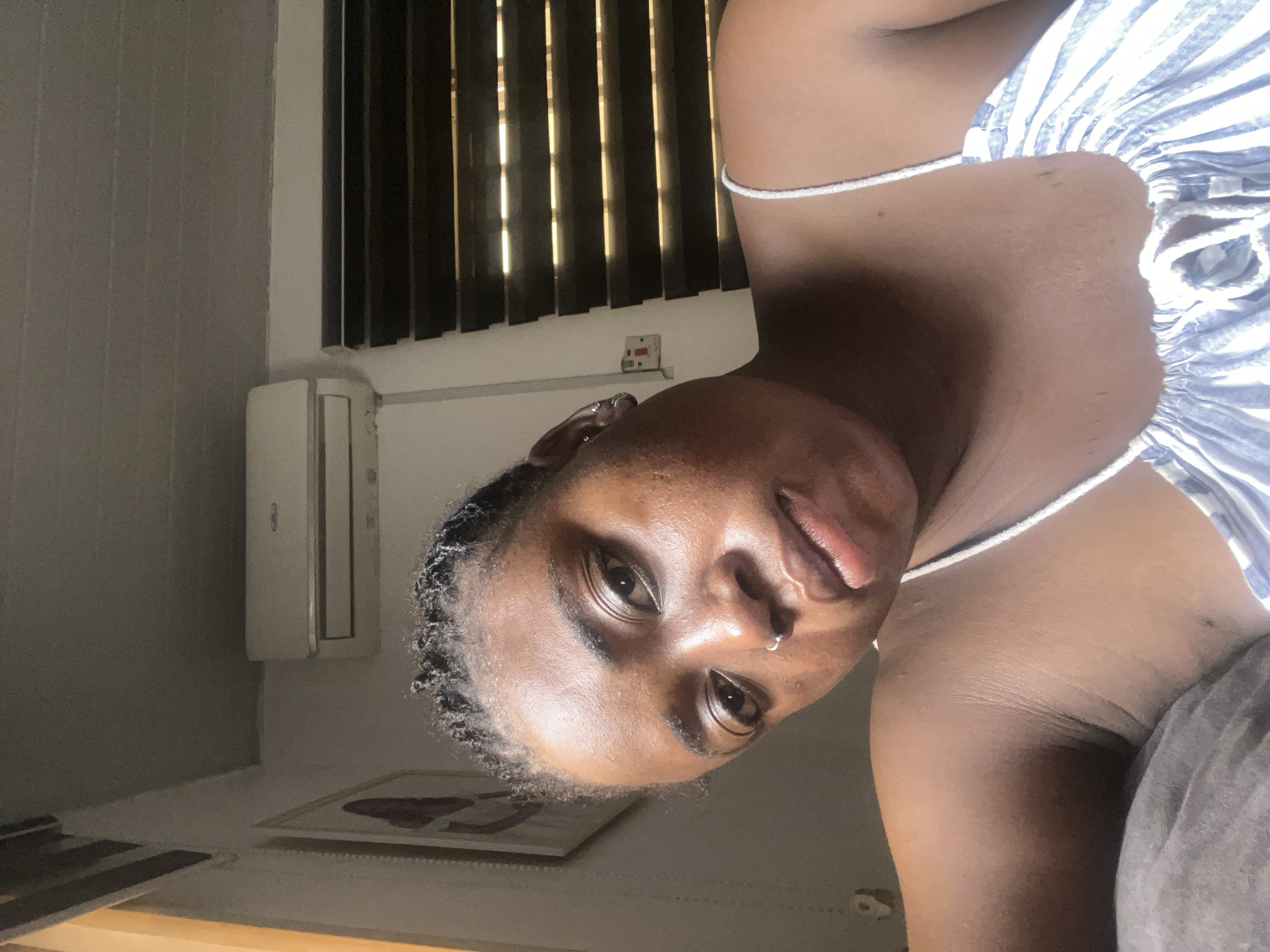 The media file features a close-up, angled shot of a young Black person, who appears to be of African descent. They are indoors, with a wall and blinds visible in the background. To the left, an air conditioning unit is mounted on the wall. In the lower portion of the image, a framed artwork is visible on a shelf or surface. The lighting suggests it might be daytime, with sunlight filtering through the blinds. The subject is looking towards the camera, with a relaxed or neutral expression. They are wearing a white and blue striped top with thin straps. A nose ring is visible on their nose. The overall ambiance is casual and personal.