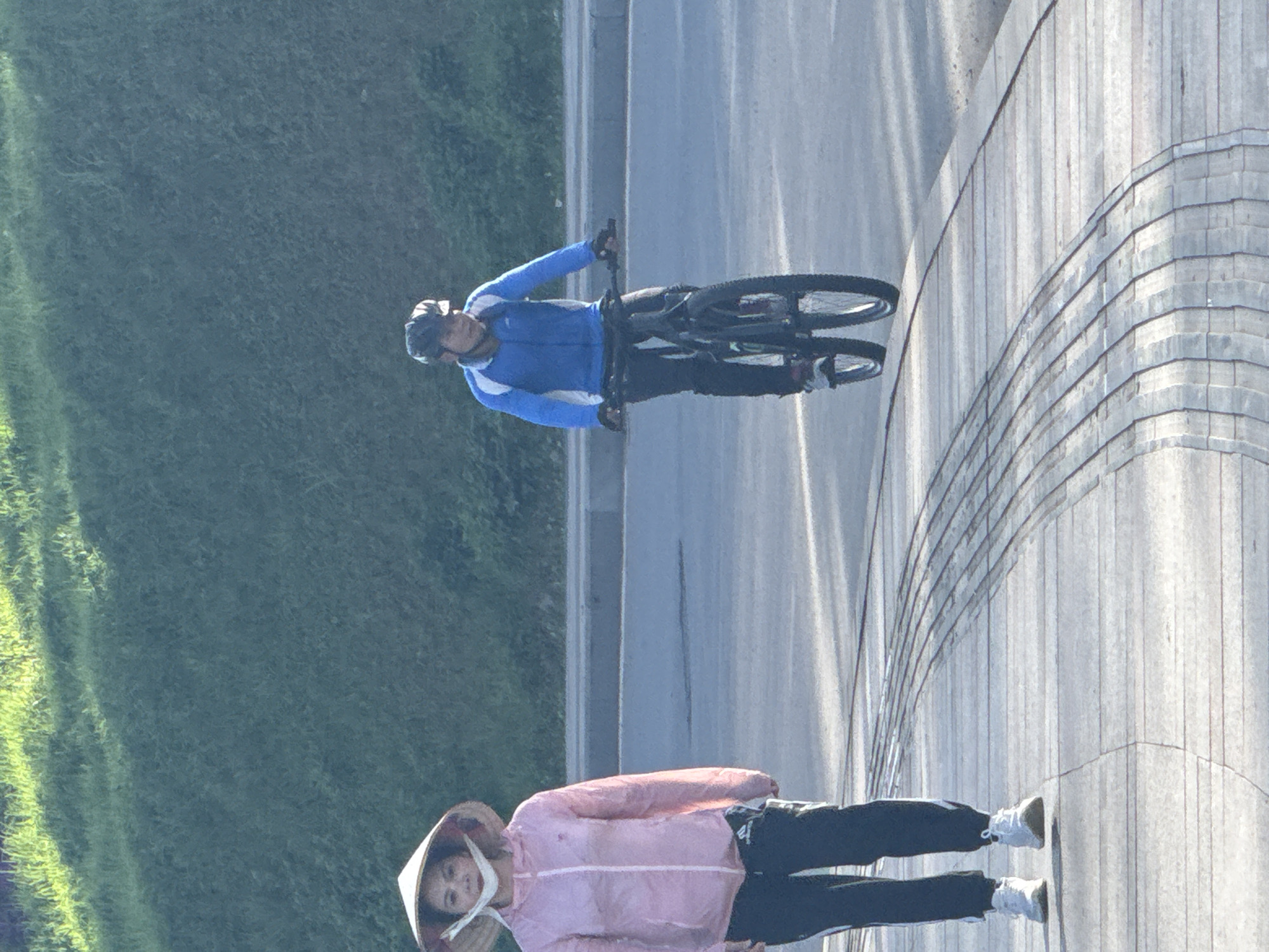A person wearing a conical hat and a pink jacket stands on a sidewalk, looking towards the right. To the right of this person, a person wearing a blue and white jacket and a helmet is riding a bicycle. The scene appears to be on a paved road with a grassy embankment in the background. The image suggests an outdoor setting in Da Lat, Vietnam, possibly during daylight hours.