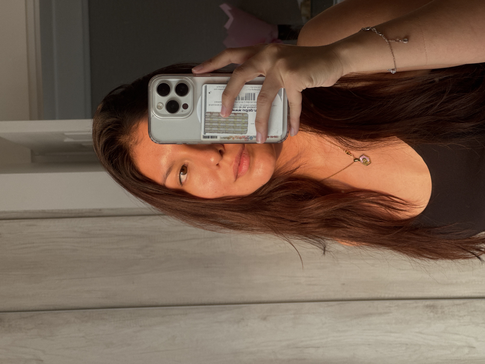 An indoor mirror selfie is captured, showing a person holding a smartphone, with the image currently rotated 90 degrees counter-clockwise. The central subject is a person, likely female, with long, straight, dark brown hair that cascades over their shoulder. Their face is partially obscured by the phone, revealing a brown eye, a portion of their nose, and pink lips with a neutral expression. They wear a black top and adorn their left wrist with a delicate silver chain bracelet featuring a small charm, while a necklace with a hexagonal, possibly amber or stone, pendant hangs around their neck.
The person holds a light-colored smartphone, possibly silver or white, encased in a clear protective cover. Behind the phone, several cards or IDs are visible within the clear case.
The background is a reflection of the room where the photo is taken. A light-colored wooden surface, appearing like floorboards or wall paneling, dominates the lower half of the reflection. In the upper reflected background, a soft pink object (perhaps a flower arrangement or decoration) is faintly visible, along with darker and lighter wall areas, suggesting an interior space. The lighting is warm and directional, casting a golden hue on the person's skin and hair, indicative of late afternoon or early evening light.
Visible text on the cards behind the phone, which appears mirrored and upside down due to the reflection and OCR processing, includes fragments like "НИЕ", "EX HYCIGUQY 29", "Jumno", "n.qslbม.www", "onema asl eb bsb", "20 niz Abel 100 05055", "GERST", and "boursgou". This scene is set in an indoor location within San Francisco Ocotlán (Ocotlán), Mexico.