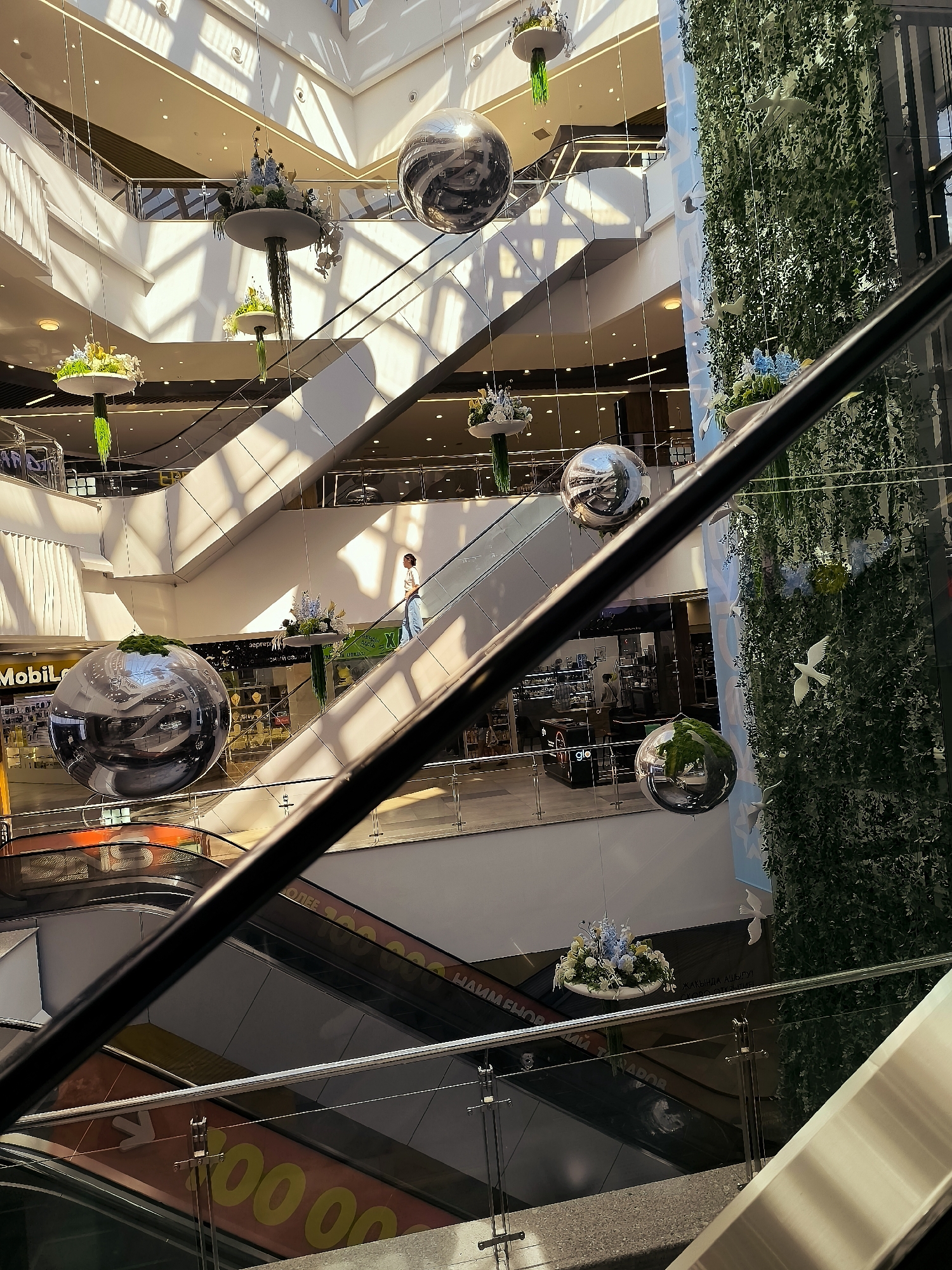 The image shows the interior of a multi-level shopping mall, likely in Qarağandı city administration, Kazakhstan, based on provided context. Sunlight streams in from a large skylight, casting diagonal shadows across the escalators and walkways. Decorative elements, including large reflective spheres and hanging floral arrangements, are suspended throughout the atrium. Greenery covers one of the walls, with white bird-shaped ornaments attached to it. Several escalators are visible, some in use with people ascending. The lower levels of the mall feature shops and commercial spaces, with large banners visible on the escalators displaying text and numerals. A person is seen walking on an escalator in the middle ground. The foreground is partially obscured by a dark railing and a portion of an escalator.