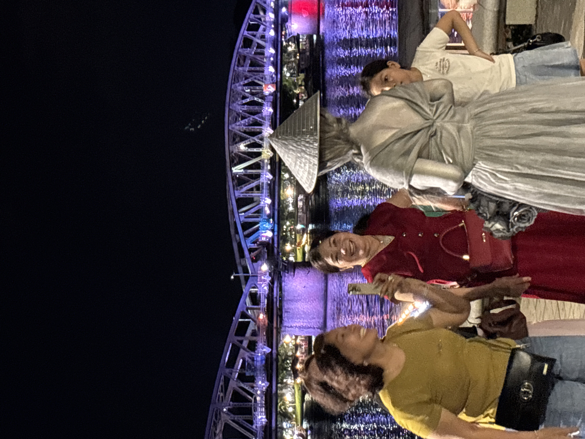 The image shows a group of people standing on a walkway next to a brightly lit bridge at night. The bridge, illuminated with blue and purple lights, spans across a body of water reflecting the vibrant colors. Several individuals are captured in the frame. In the foreground, two women are smiling and looking towards the camera, with one holding a phone. One woman is wearing a yellow t-shirt and jeans, and the other is wearing a red dress. Behind them, a person in a white t-shirt and blue jeans is also visible, with a person in a silver dress and a conical hat standing close by. The scene appears to be a festive or tourist area, likely in Thuận Hóa Ward, Vietnam, given the presence of the conical hat and the distinctive bridge lighting, which resembles the Dragon Bridge in Da Nang. The overall mood is cheerful, with people posing for photos. The time of day is clearly night, evidenced by the dark sky and artificial lighting.
