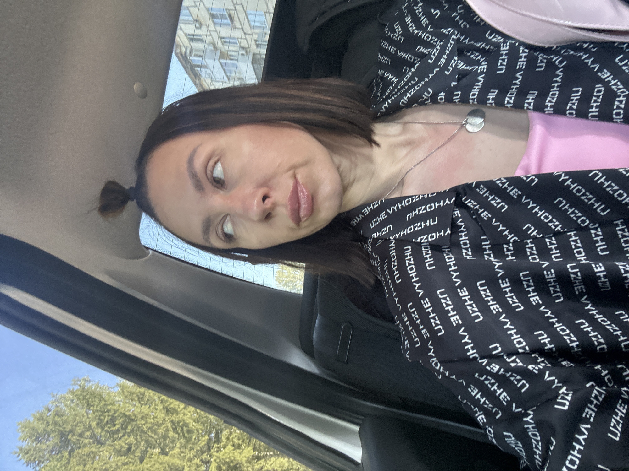 A woman with short brown hair partially tied up is sitting in a vehicle, wearing a black jacket with white lettering, and a pink top underneath. She is looking to the left with a slight smirk. A silver necklace with a round pendant is visible around her neck. Outside the vehicle window, green trees and a building can be seen under a clear, blue sky. The interior of the vehicle is visible, including the seat and parts of the frame around the window. There's no indication that this image was taken in Votkinsk, Russia.