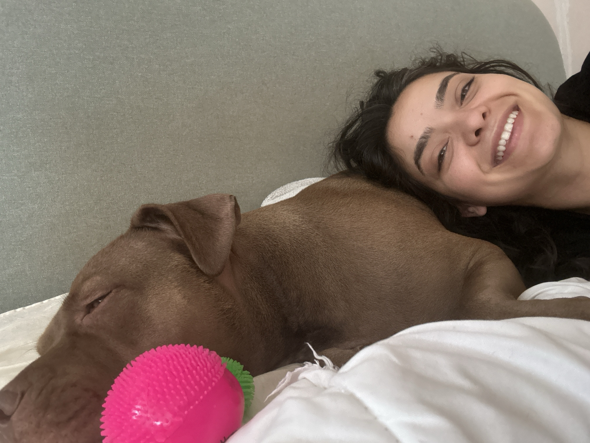 A young woman with dark hair and a wide smile rests her head on a brown dog. They are lying on a white bed, possibly in a bedroom. The dog is sleeping or resting with its eyes partially closed, and a bright pink spiky ball is positioned near its head. The background features a textured grey headboard. There is no indication that this scene is located in Malevo, Bulgaria.